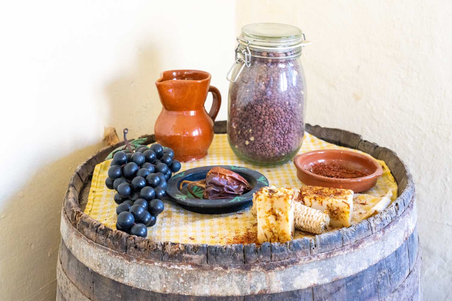 Assorted foods on a barrel: grapes, red clay jug, spices, a jar of beans, and seasoned cheese on a yellow cloth.