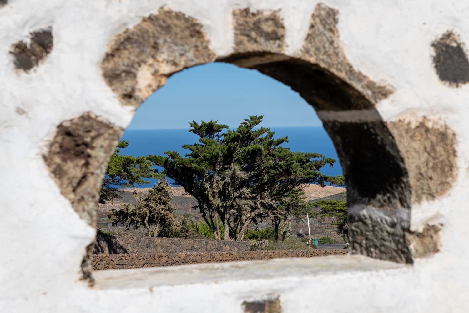 Stone wall window framing scenic view of trees and ocean horizon under clear blue sky.