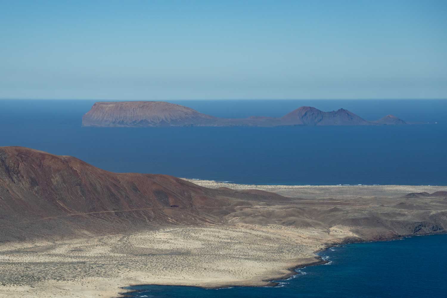 Distant brown volcanic islands with rocky coastline and deep blue ocean under clear blue sky.