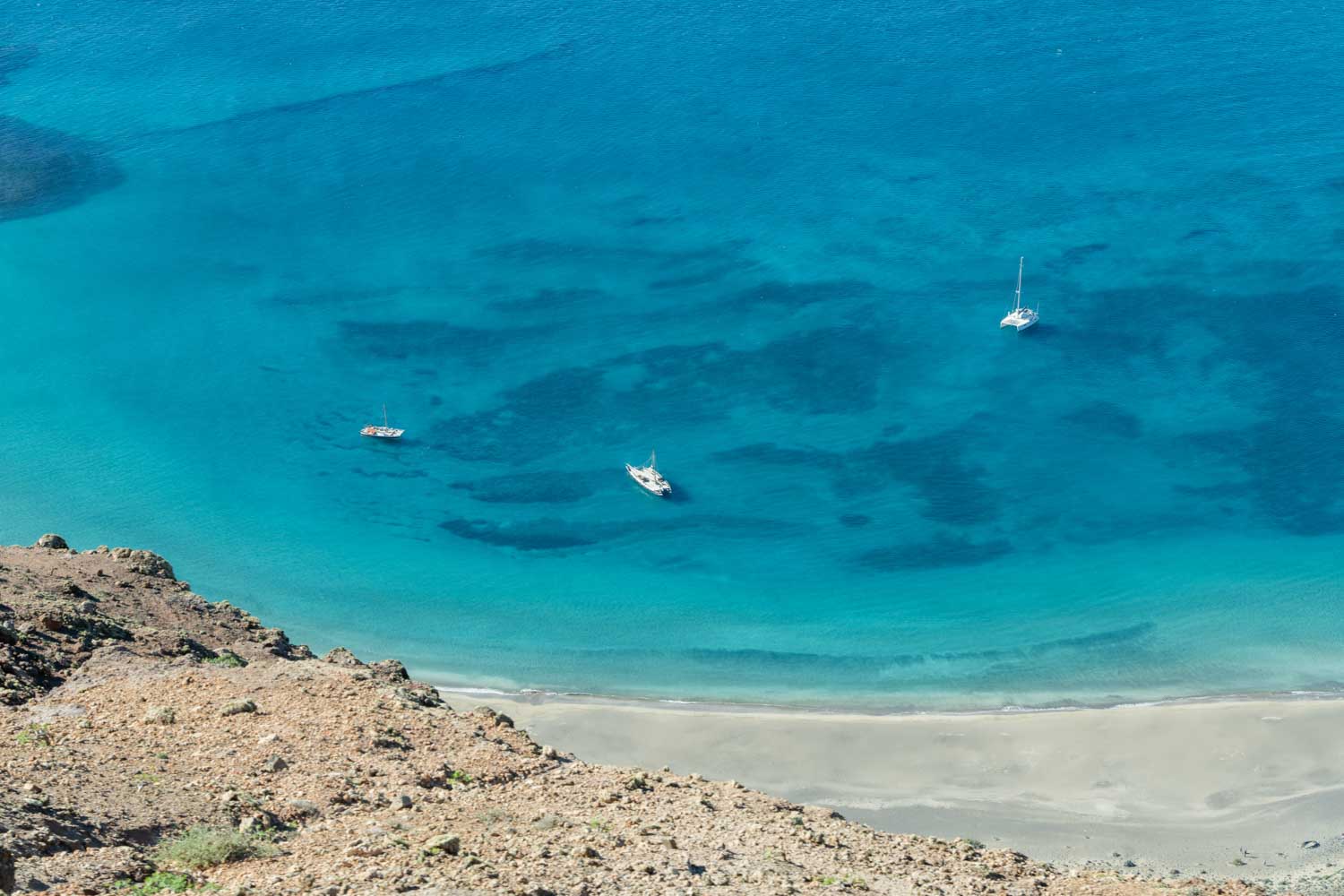 Aerial view of turquoise sea with three boats near a rocky coastline and sandy beach.