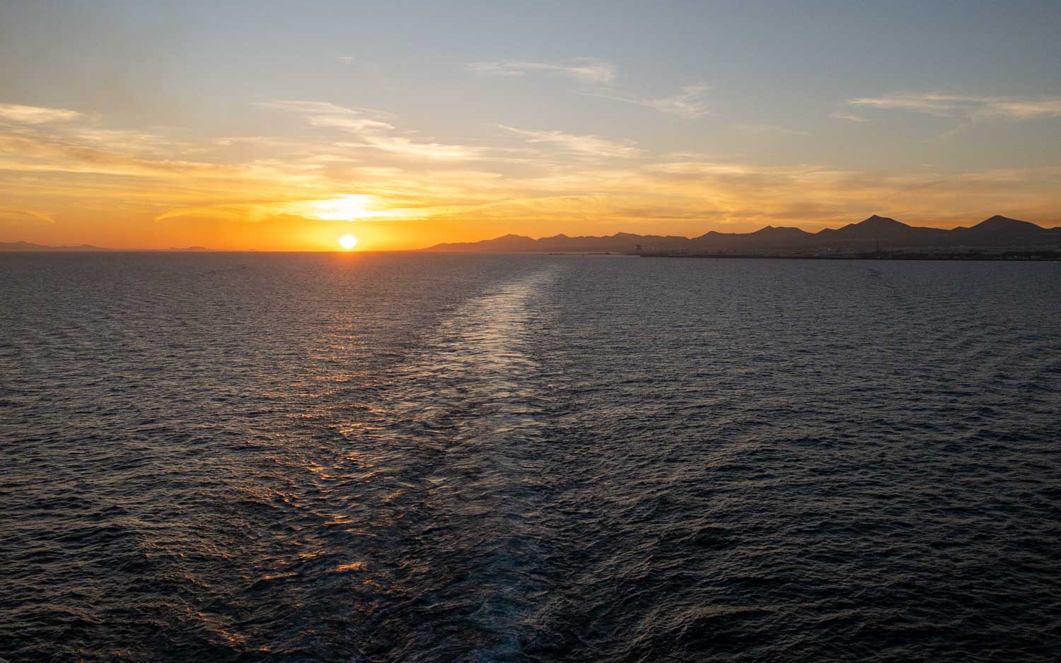 Sunset over the ocean with a visible horizon and mountains in the background, casting a warm glow on the water.