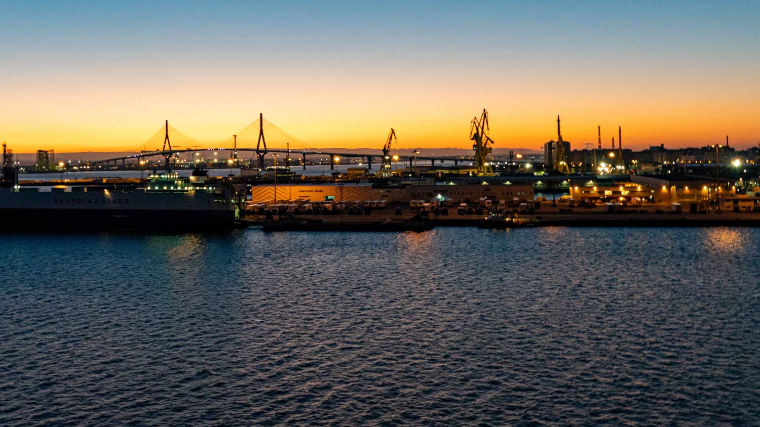 Port at sunset with illuminated cranes and distant bridge, reflecting on the water, creating a serene industrial landscape.