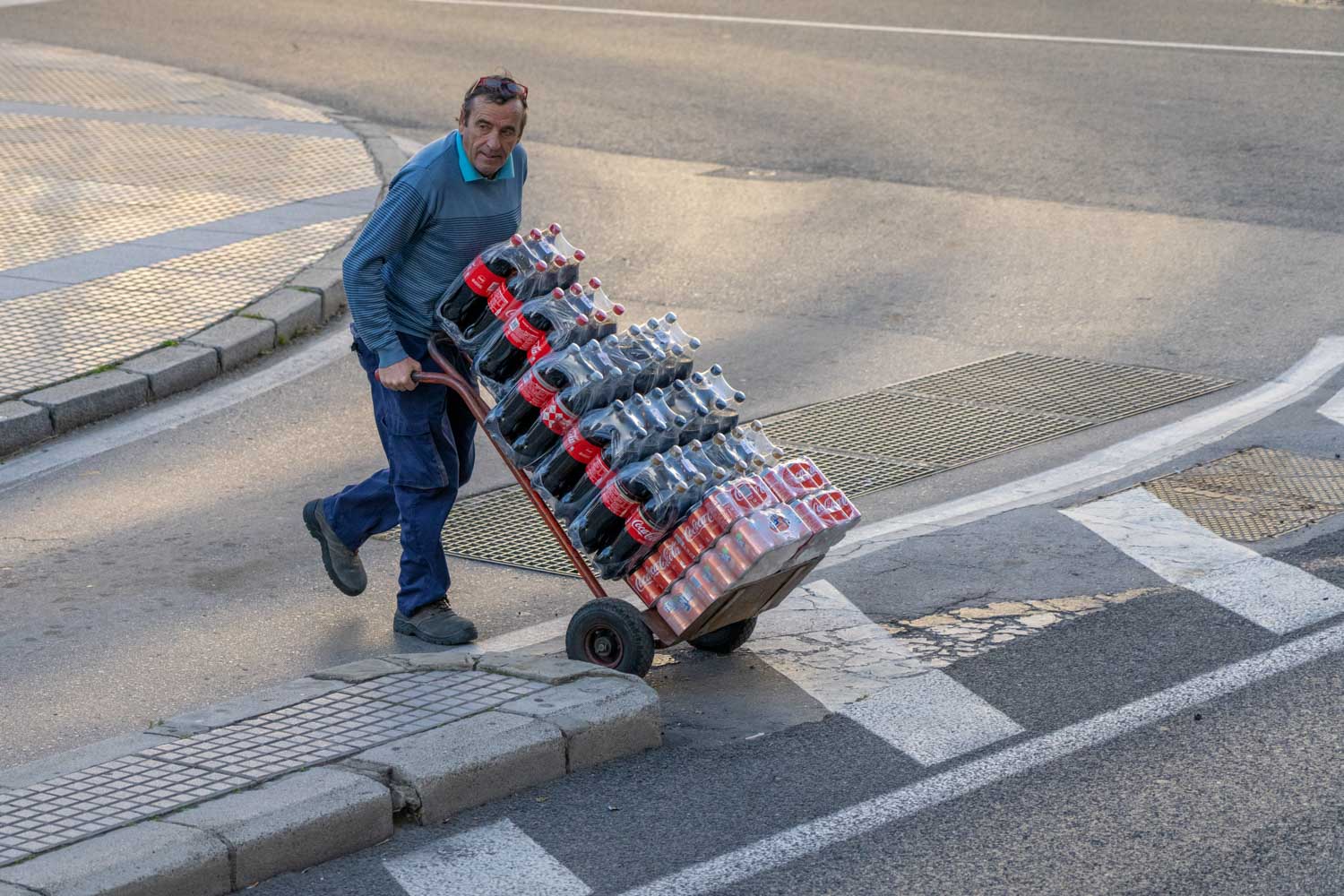 Man pushing a hand truck loaded with Coca-Cola bottles on a city street corner.