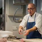 Elderly fishmonger wearing a blue apron gestures while explaining fish cuts in a market setting.