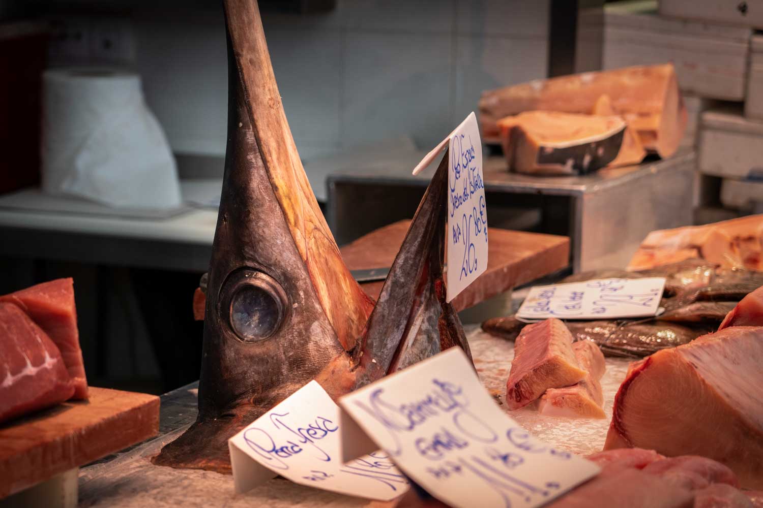 Swordfish head displayed at a fish market with price tags, surrounded by various fish cuts on ice.