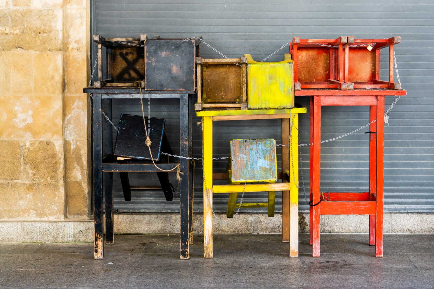 Stacked colorful wooden tables and chairs against a wall, featuring black, yellow, and red hues.