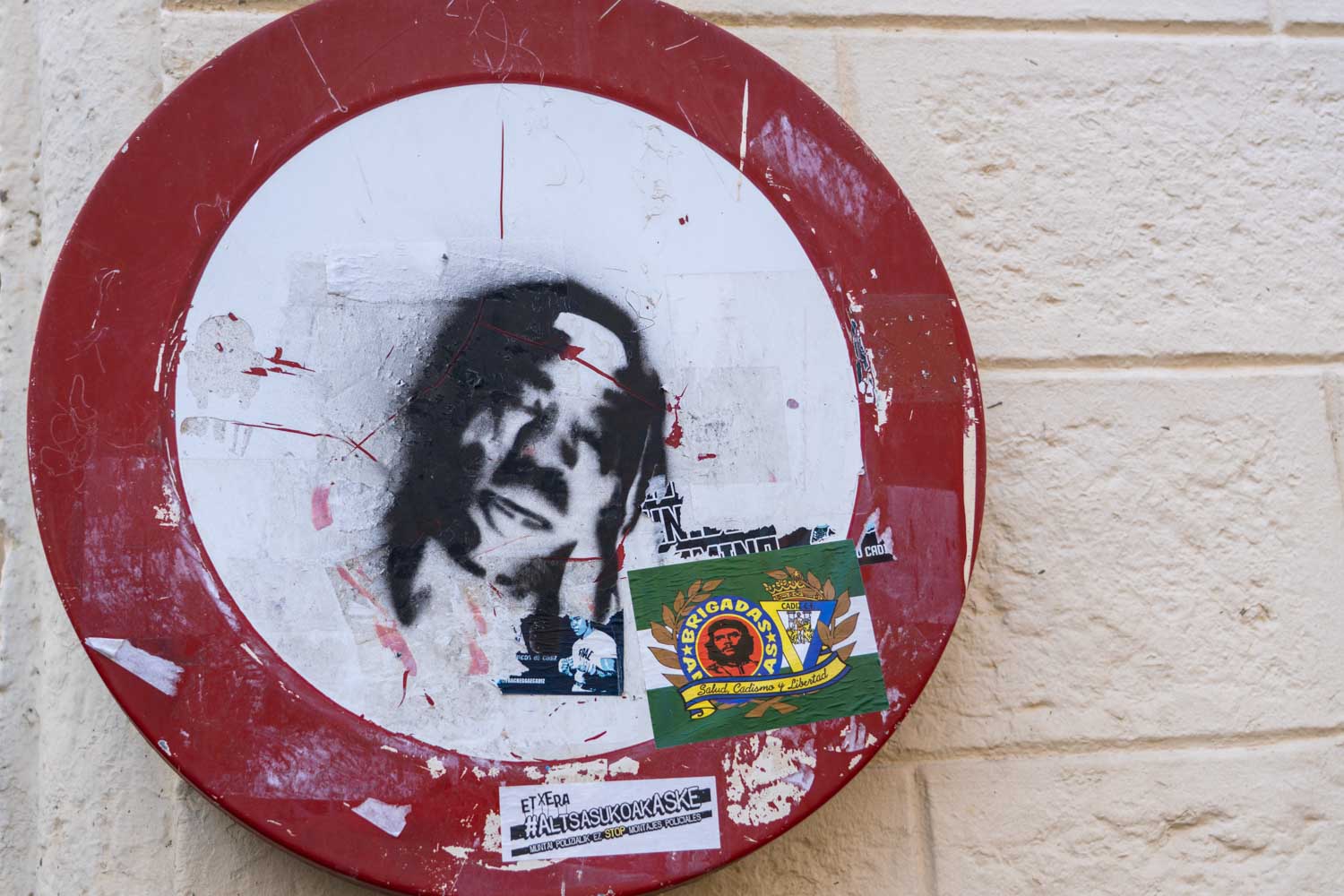Graffiti face on a defaced red and white sign with stickers on a brick wall background.