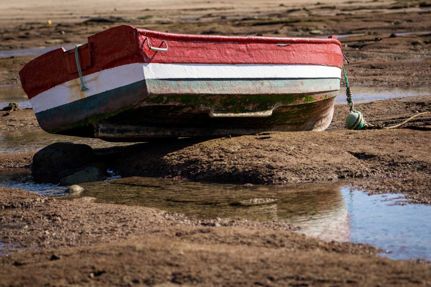 Red and white rowboat ashore on brown muddy beach at low tide, with blue sky reflection in shallow water.