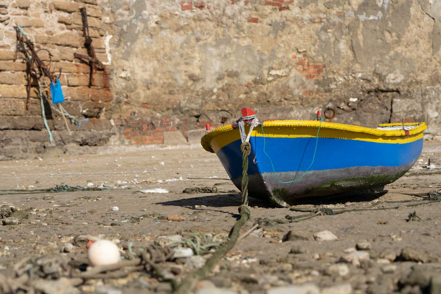 A blue and yellow boat on dry land against a weathered brick wall in low tide.