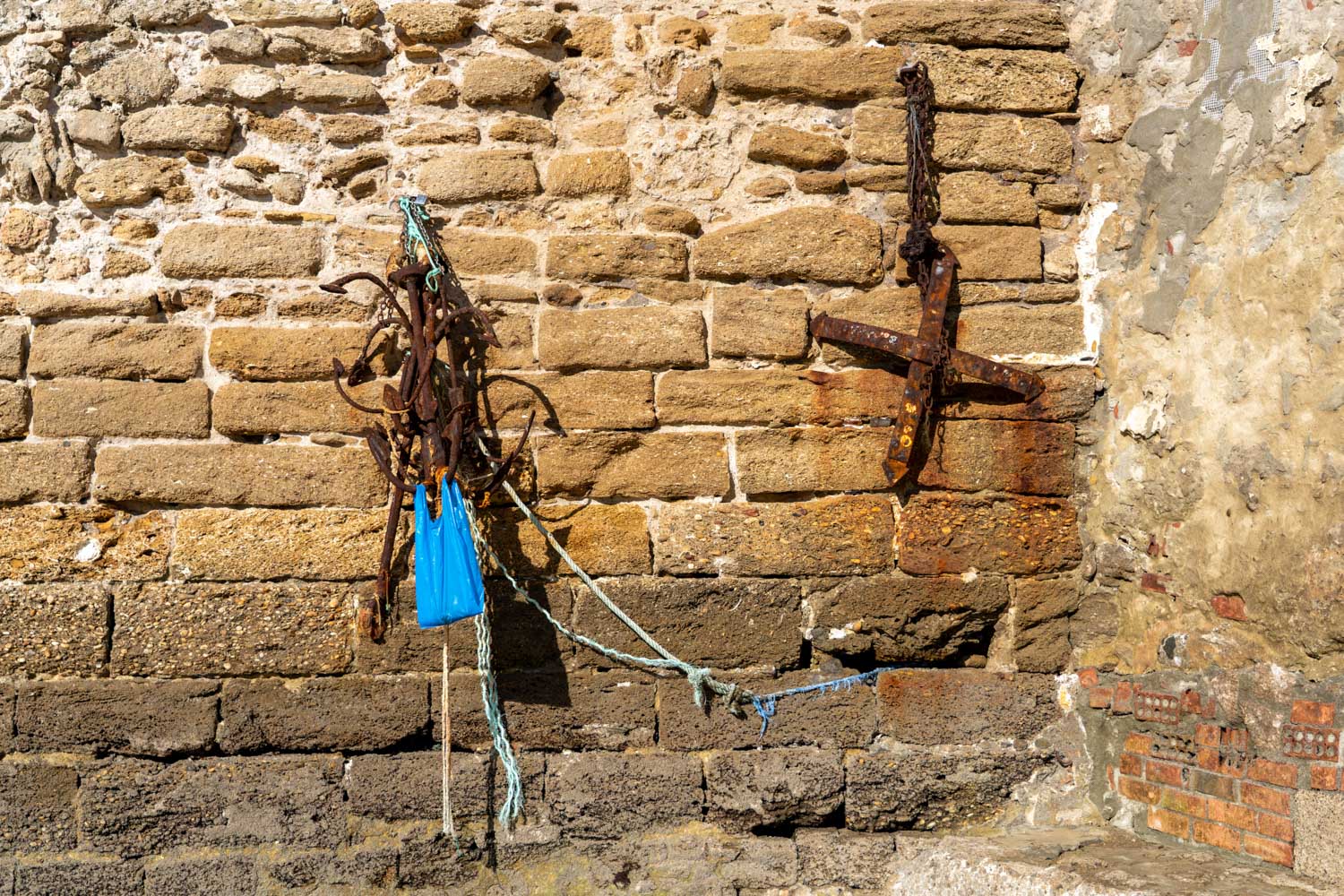 Rusted anchors with ropes against a weathered stone wall, featuring a bright blue plastic bag.