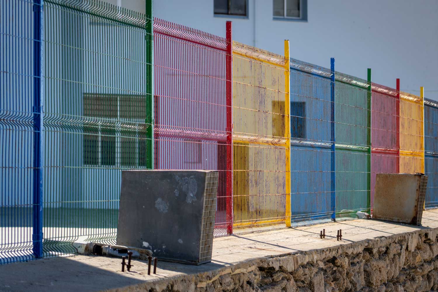 Colorful wire fence with blue, red, yellow, and green panels on a stone wall beside a building.