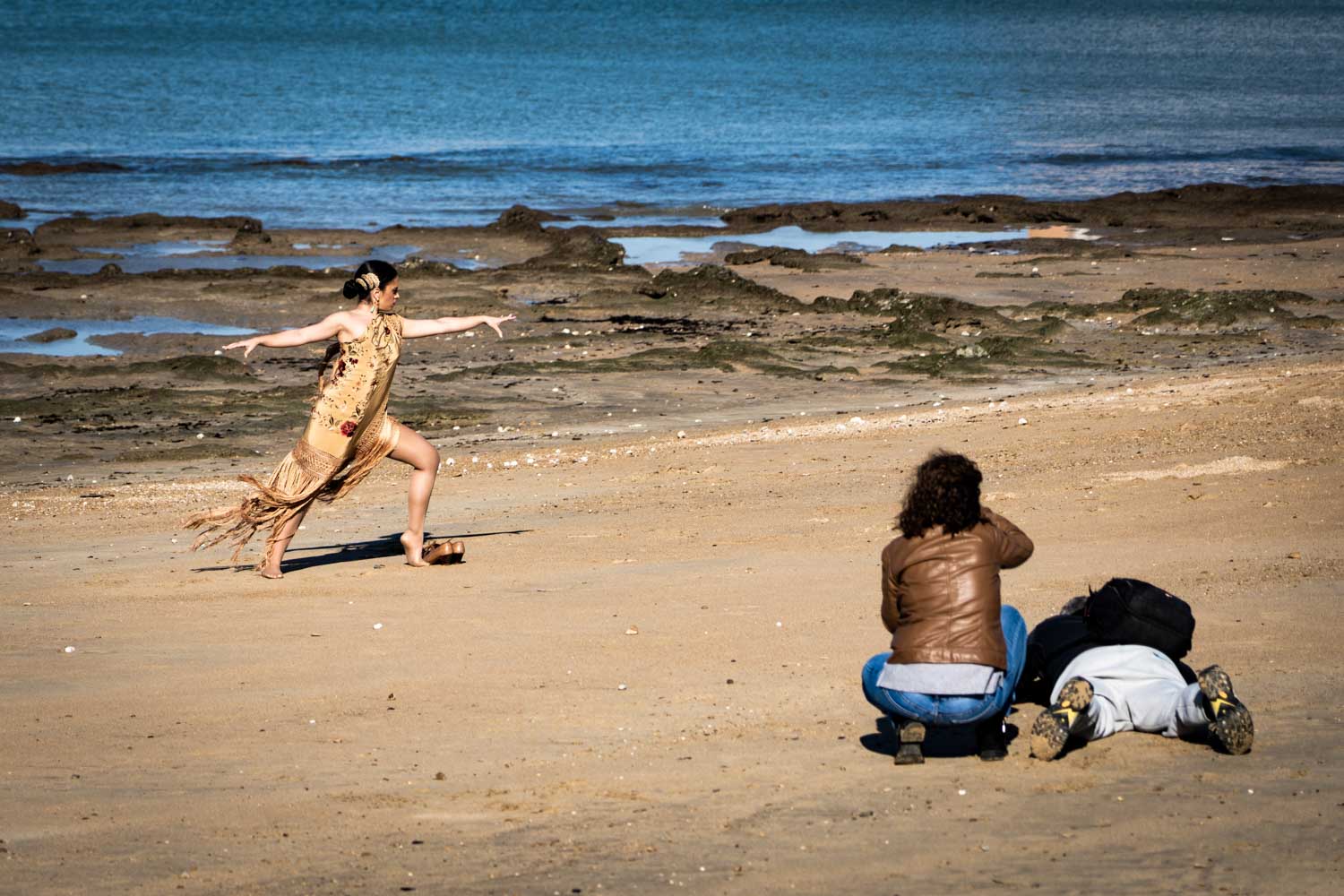 Dancer in fringed dress performs on a sandy beach as photographers capture the moment, with ocean in the background.