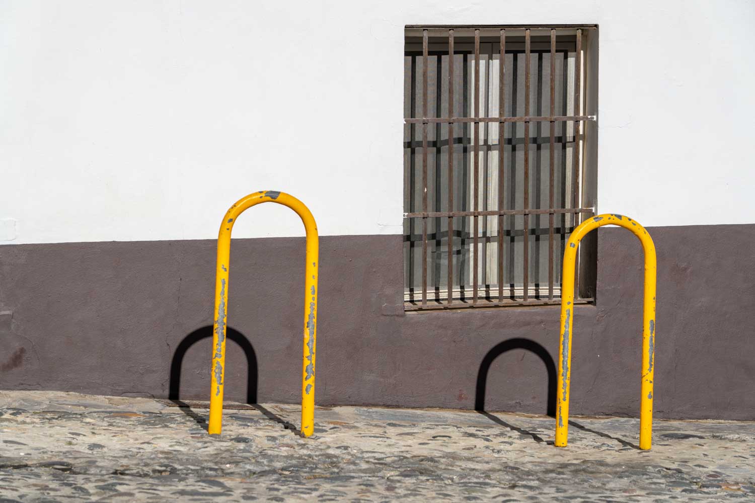 Yellow bike racks by a window with bars, set against a white and brown wall on a cobblestone street.