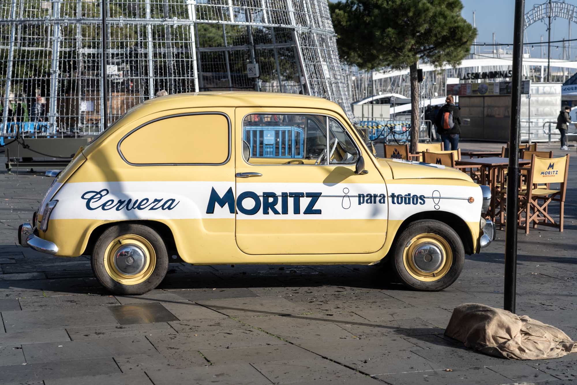 Yellow vintage car with Cerveza Moritz para todos parked outdoors, surrounded by outdoor seating and a Christmas structure.