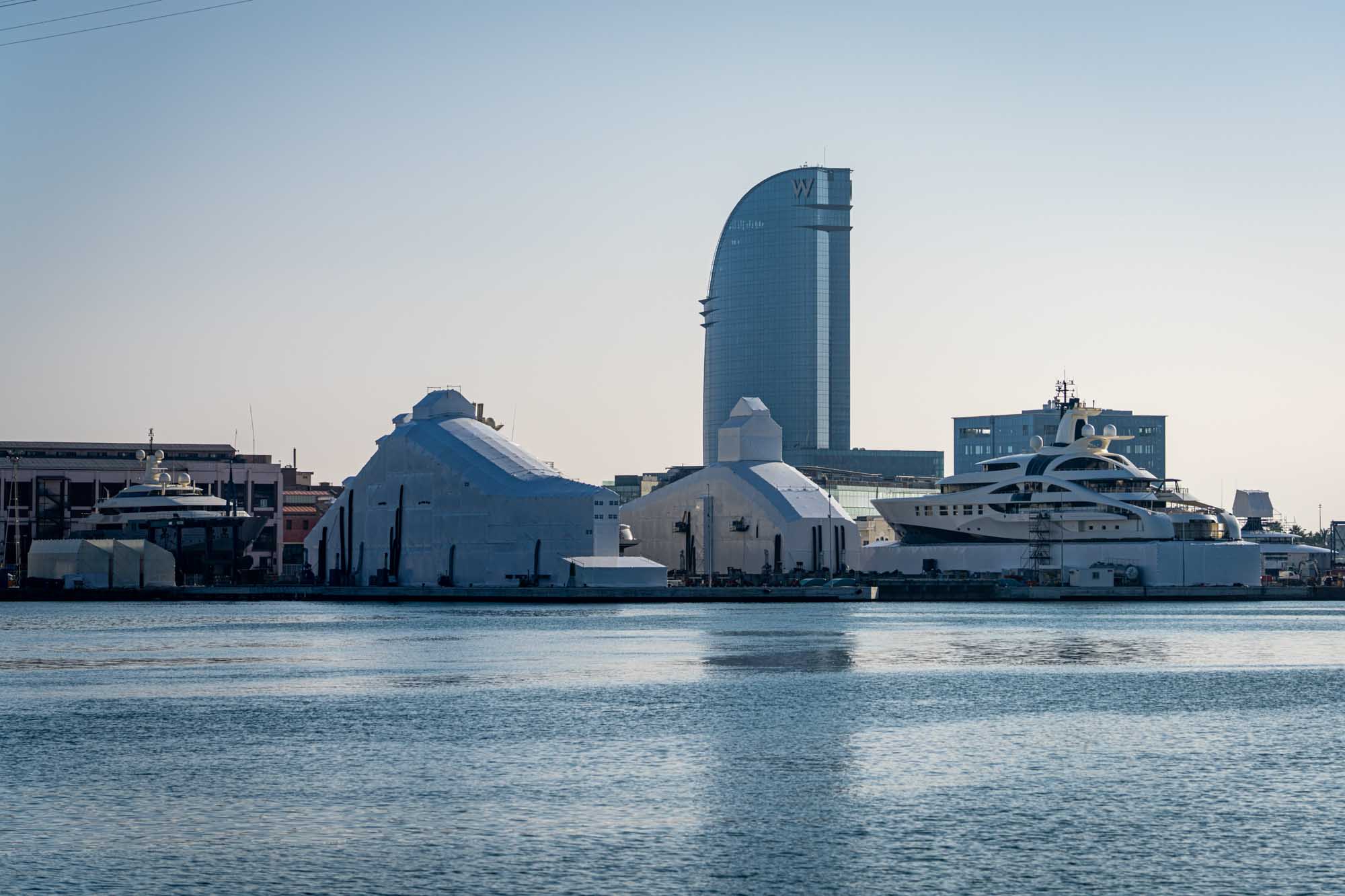 Modern waterfront with luxury yachts and a tall glass building in the background against a clear sky.