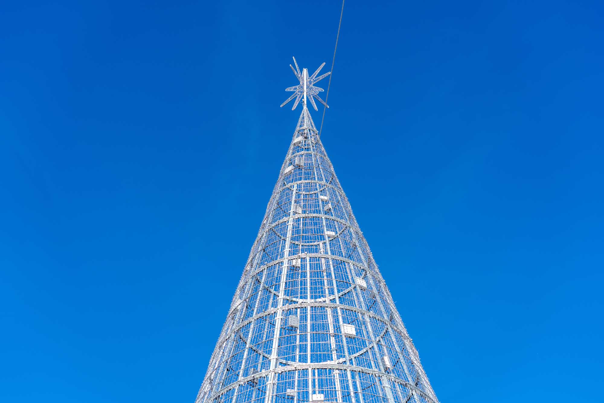 Modern metallic Christmas tree sculpture against a clear blue sky, viewed from below, featuring a star topper.