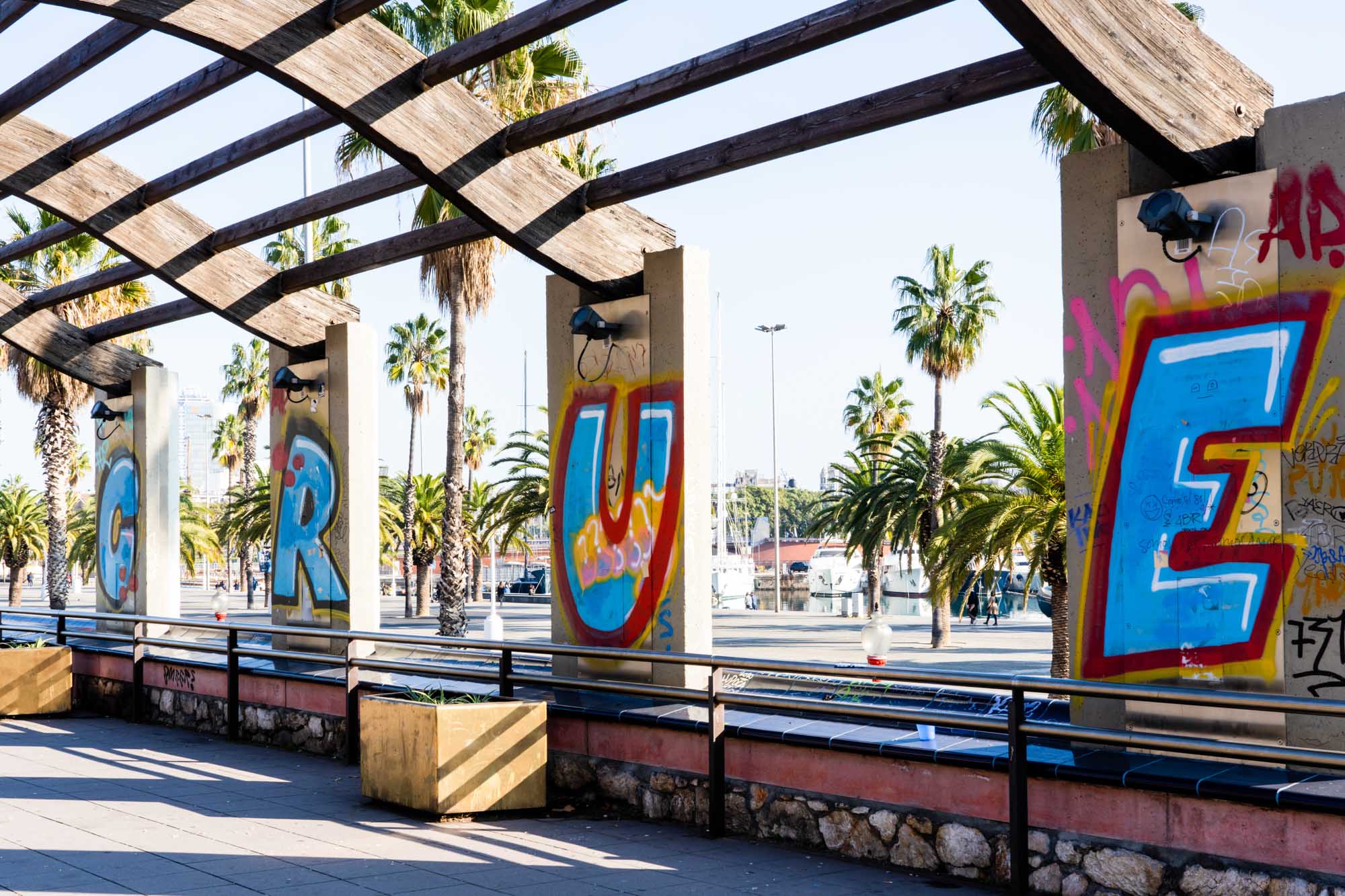 Graffiti on concrete pillars with palm trees and marina in the background, under a wooden canopy structure in sunny weather.