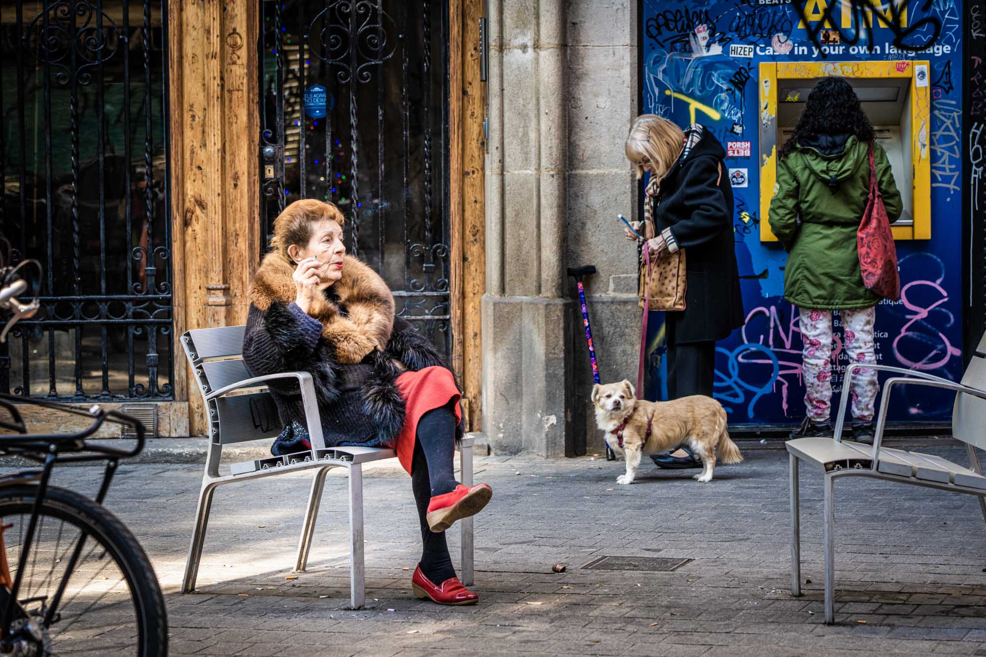 Woman in fur coat smoking on a bench, others at graffiti-covered ATM, dog on leash nearby.
