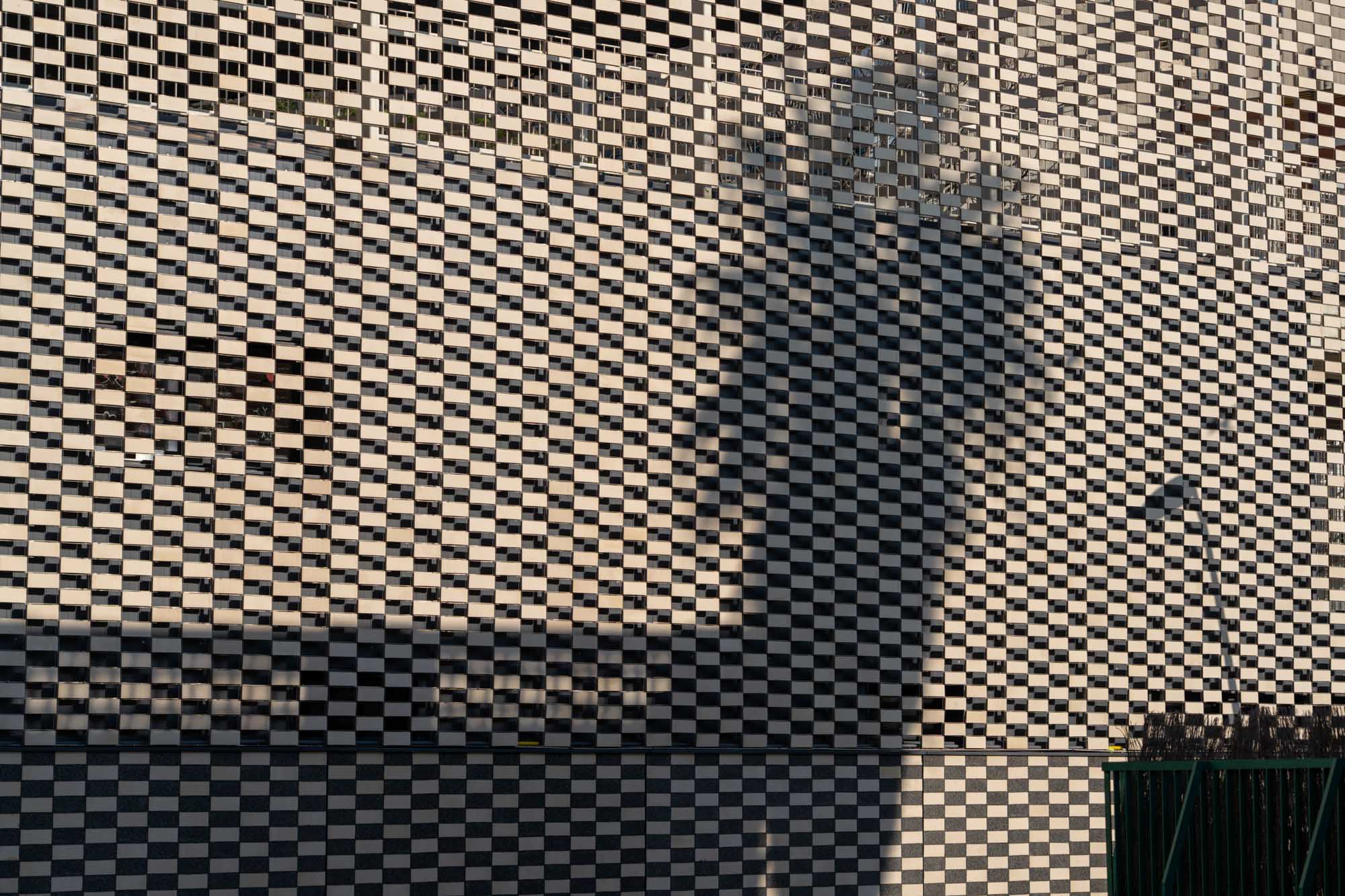 Checkerboard building facade with palm tree shadow and streetlight.