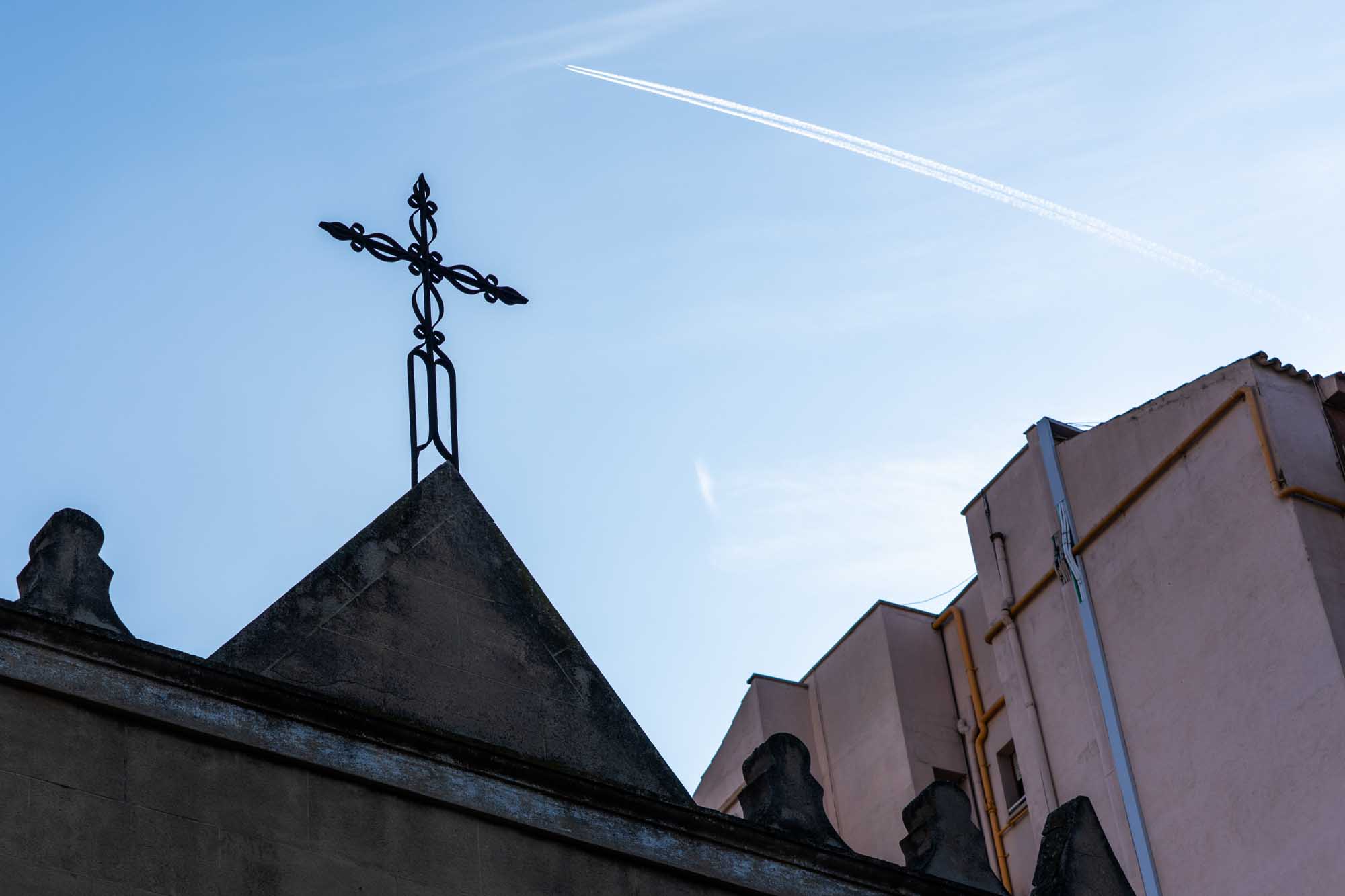 Church roof silhouette with wrought iron cross, airplane contrail in blue sky.
