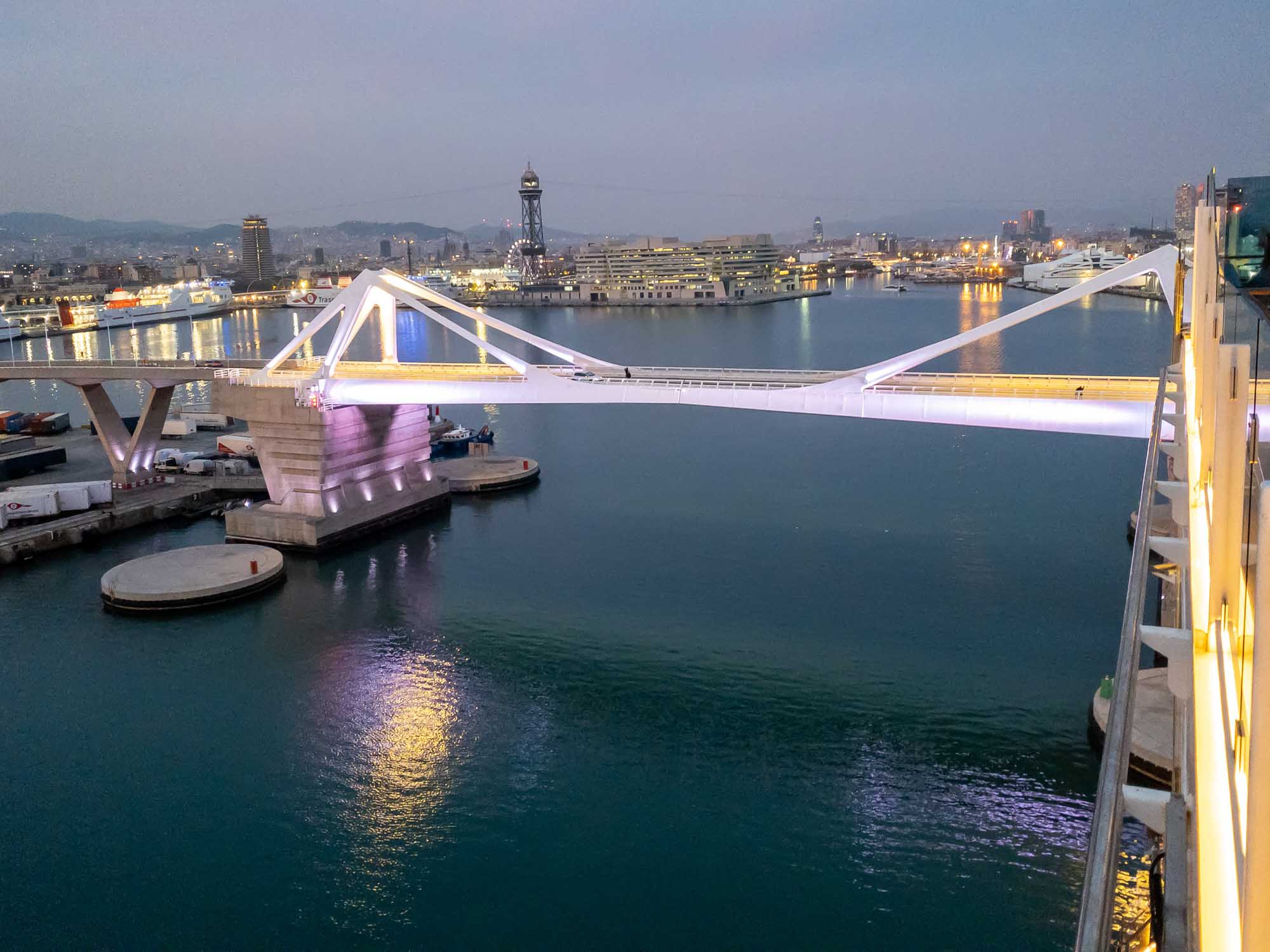 Modern bridge illuminated at dusk over a harbor with city skyline in the background.