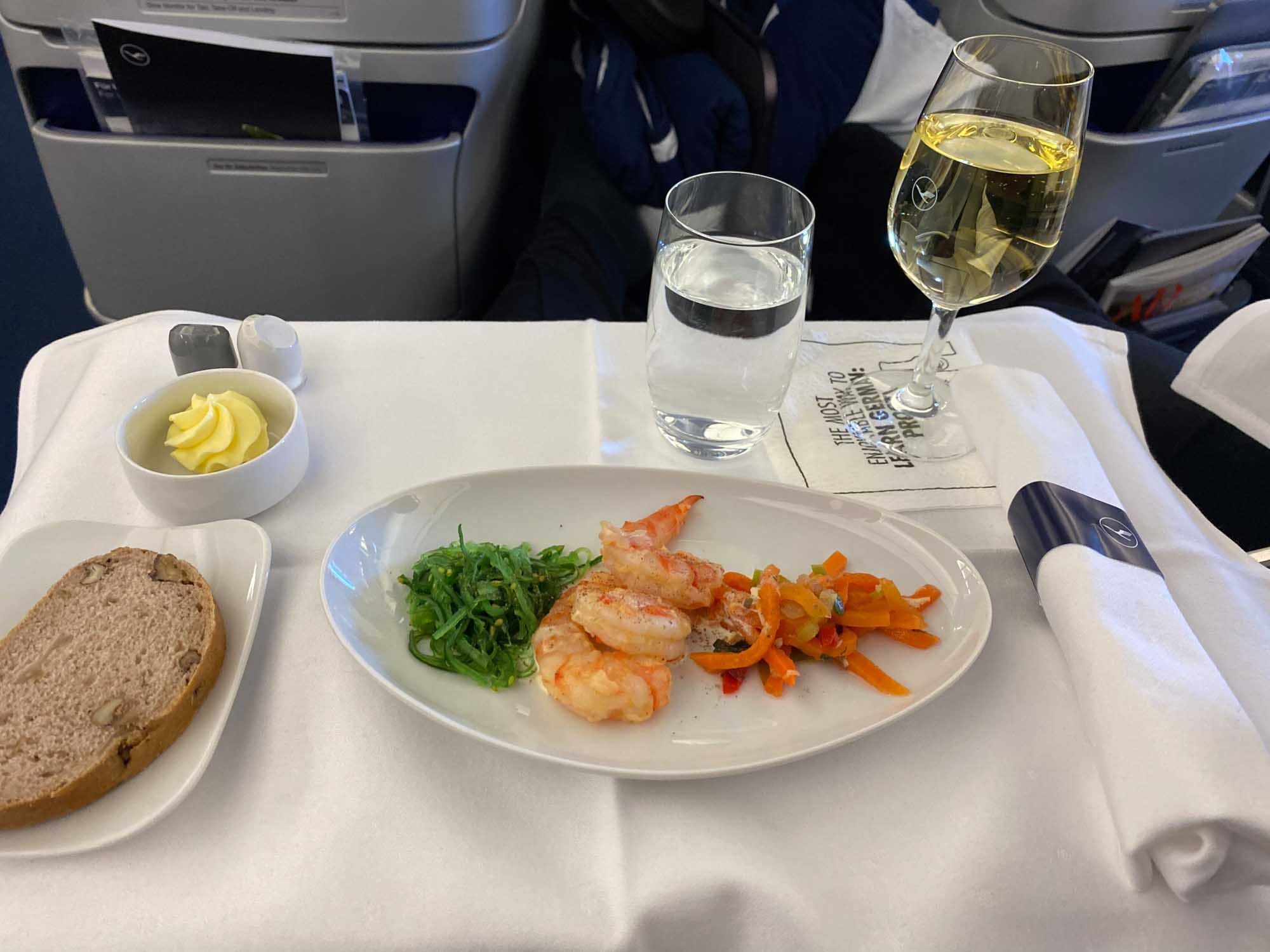Airplane meal with shrimp, seaweed salad, sauteed vegetables, bread, butter, water, and white wine in an elegant dish setting.