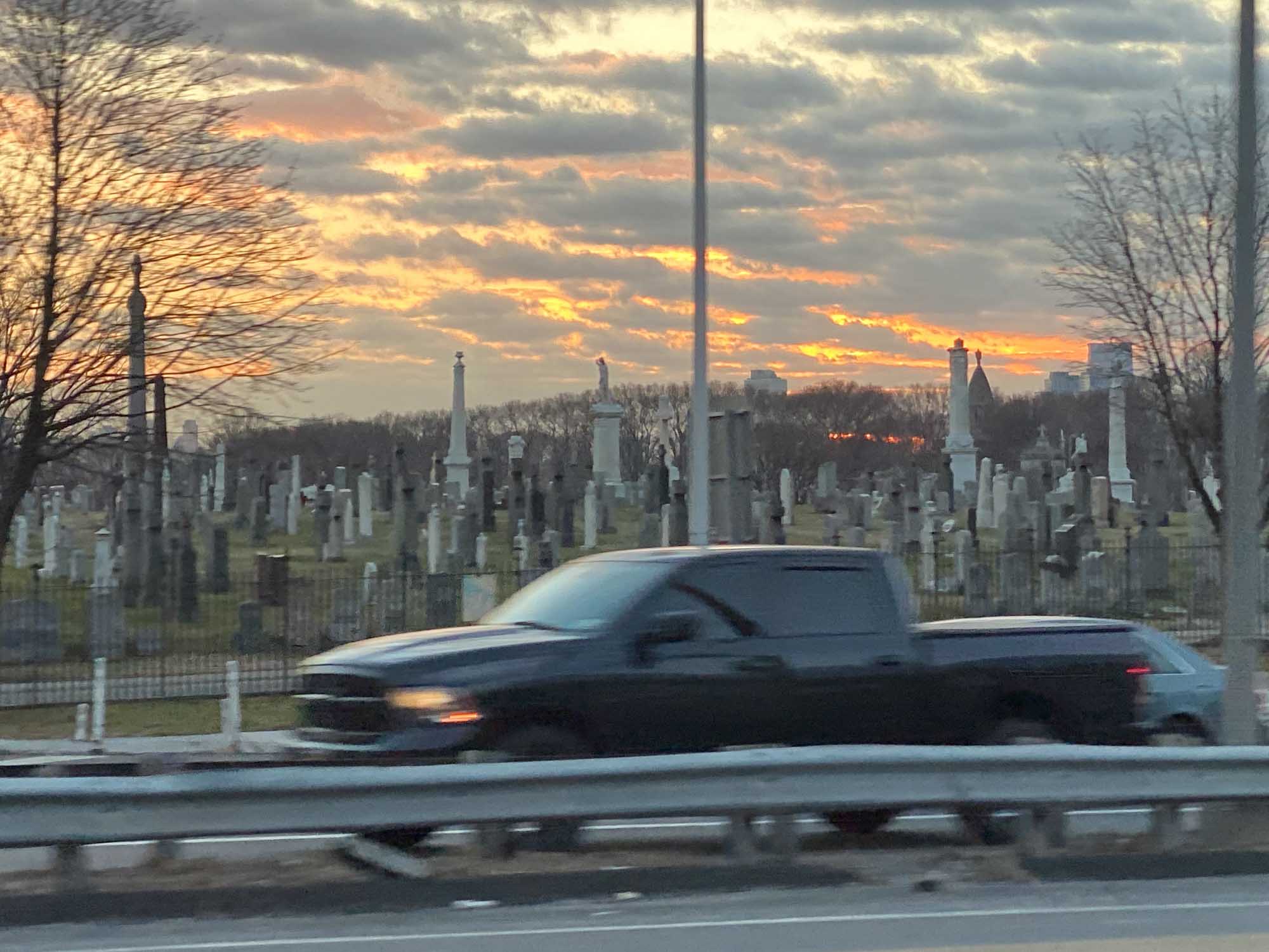Truck passing by cemetery at sunset with cloudy sky in the background.
