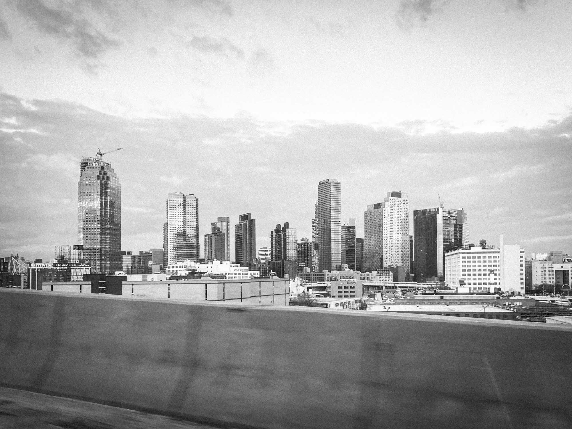 Black and white cityscape featuring tall buildings and cloudy sky viewed from a highway.