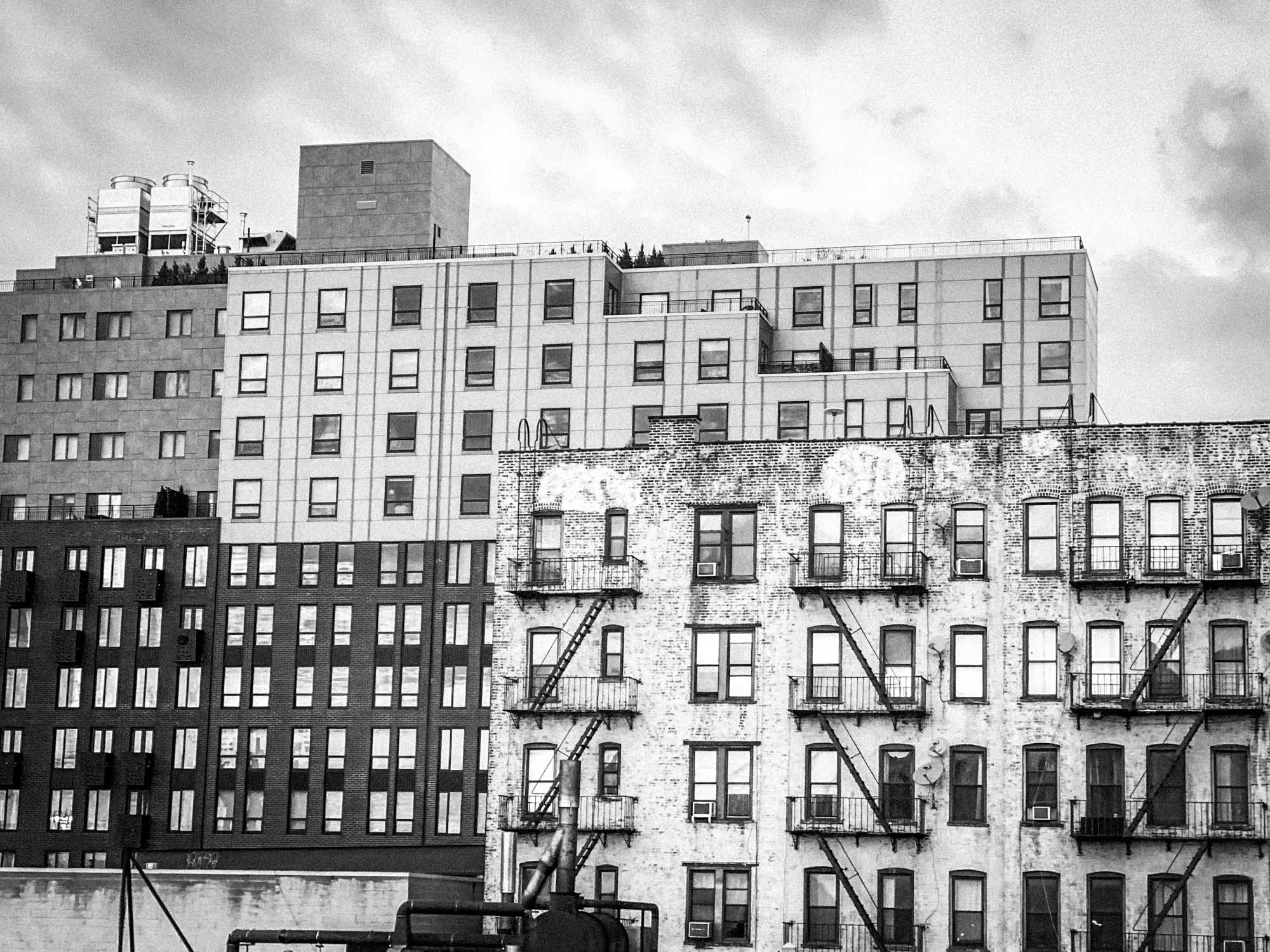 Black and white photo of urban buildings with fire escapes and rooftop water tanks under a cloudy sky.