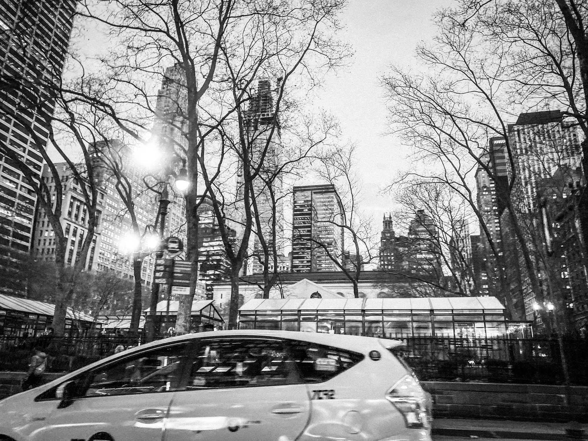 Monochrome city scene with skyscrapers, leafless trees, and moving white car at dusk. Bright streetlights illuminate the area.