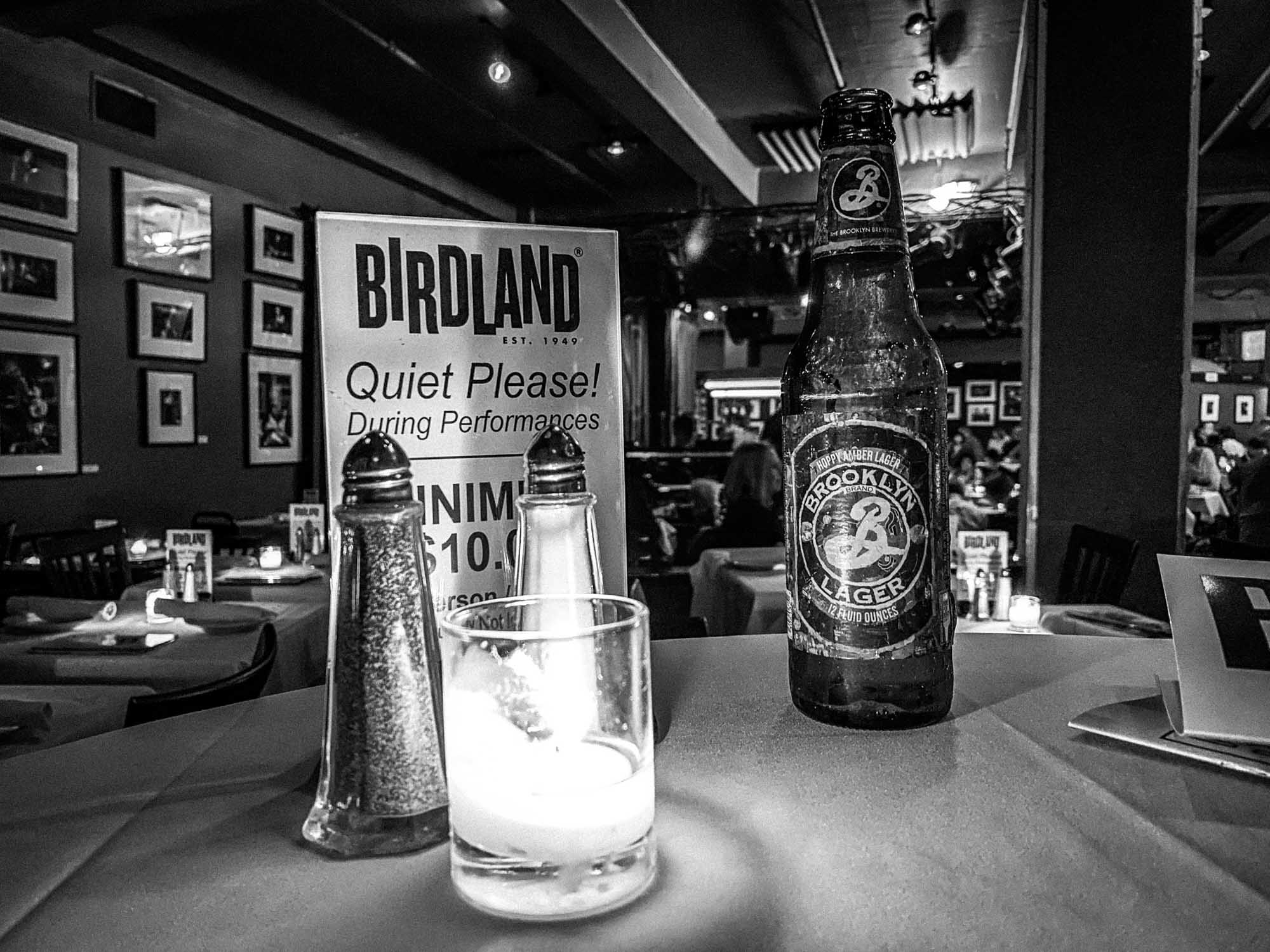 Brooklyn Lager bottle on a candlelit table at Birdland jazz club, with framed photos and dining tables in the background.