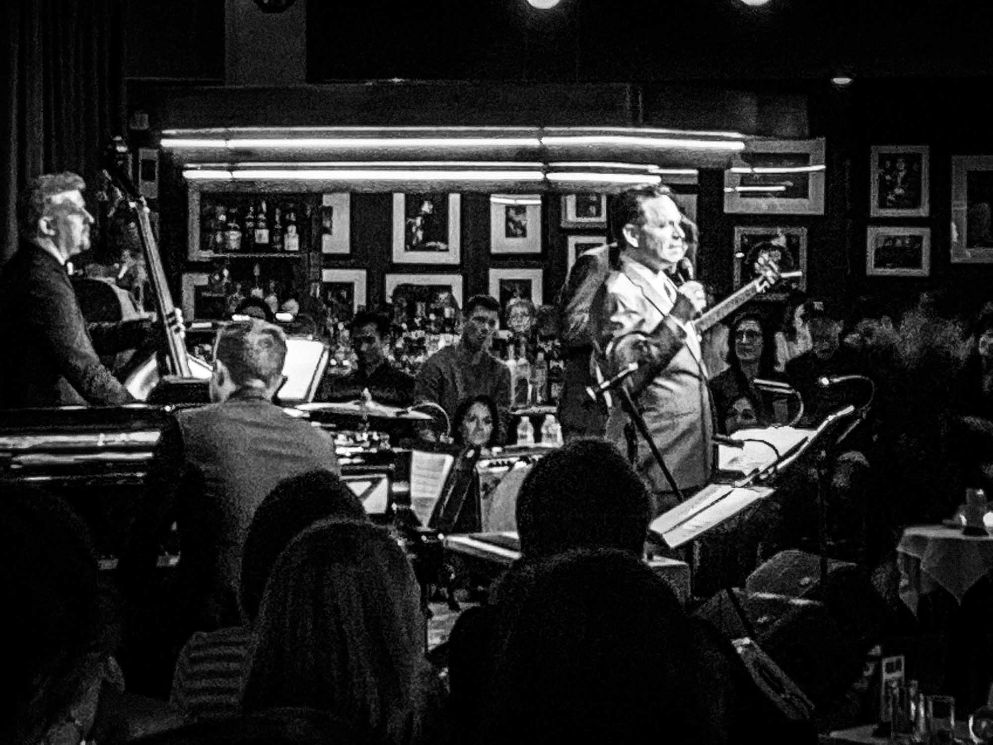 Black and white image of a jazz band performing in a cozy club, with audience engaged and musician in focus.