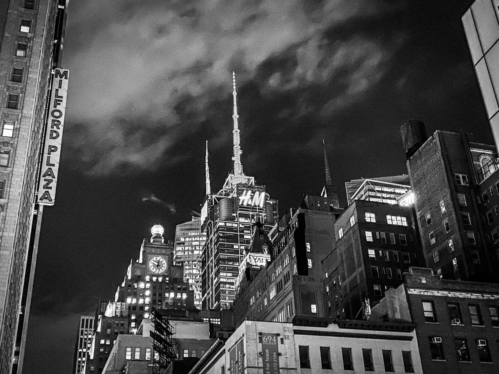 Nighttime cityscape with Milford Plaza sign and illuminated high-rise buildings under a cloudy sky.