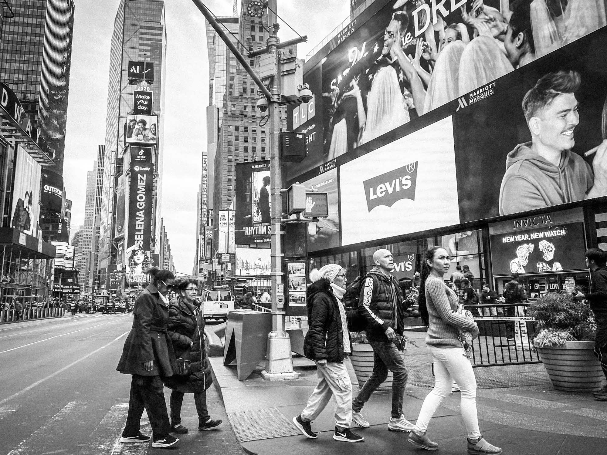 People crossing a street in Times Square, New York, surrounded by large billboards and high-rise buildings.