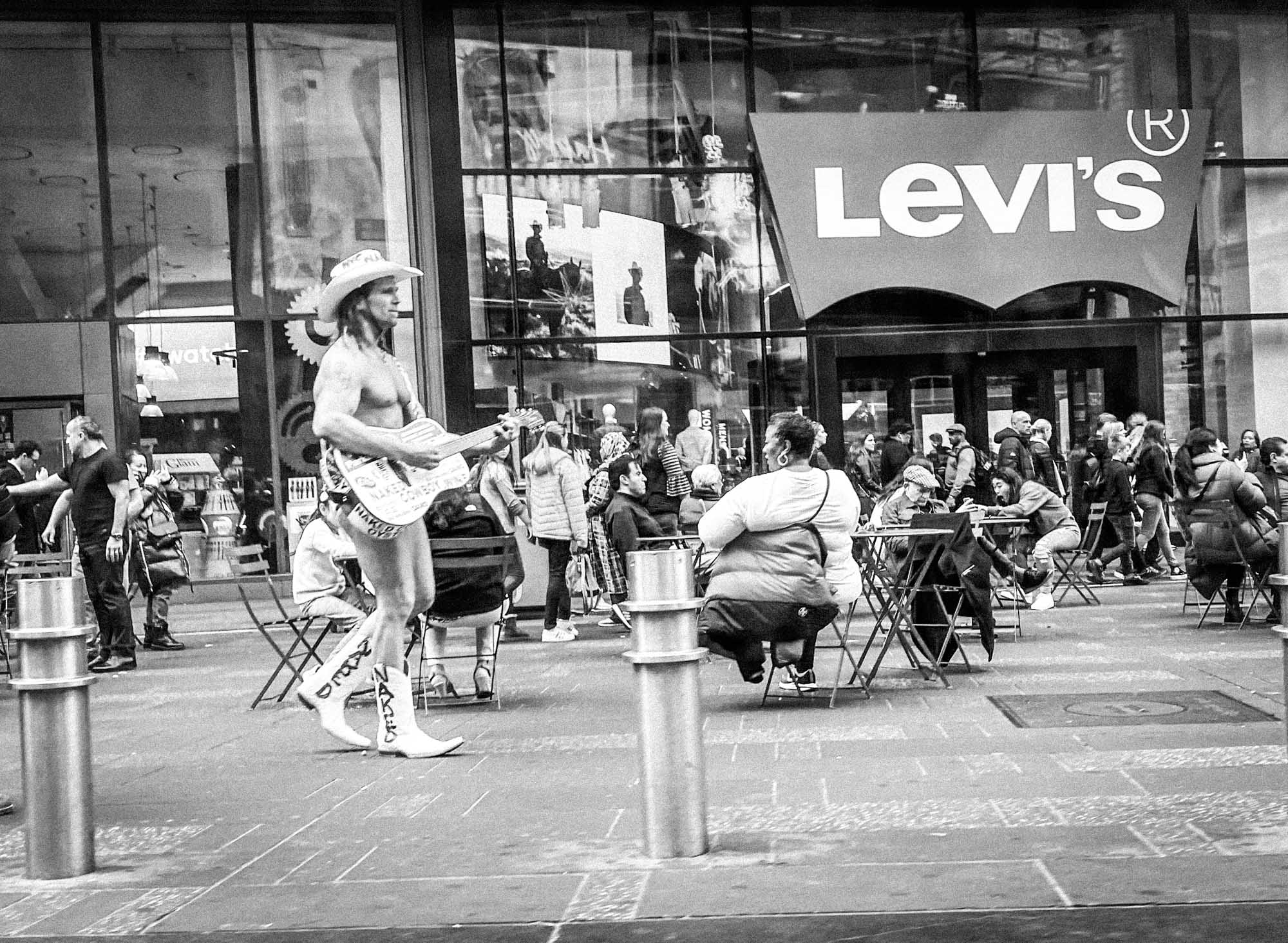 Street performer in cowboy hat playing guitar in front of Levi's store, NYC crowd sitting and walking by.
