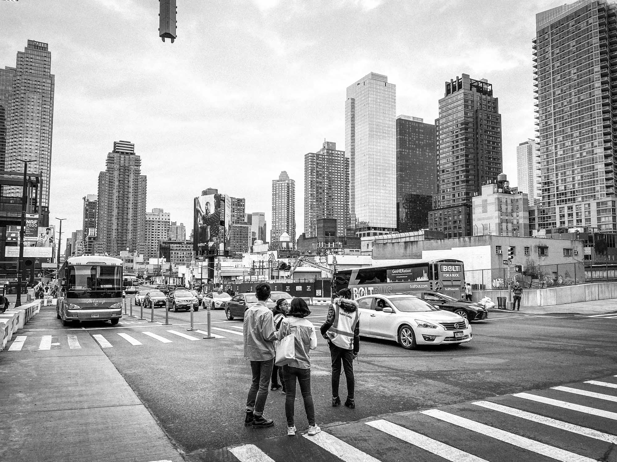 Three people crossing a busy city street with skyscrapers and traffic in the background on an overcast day.
