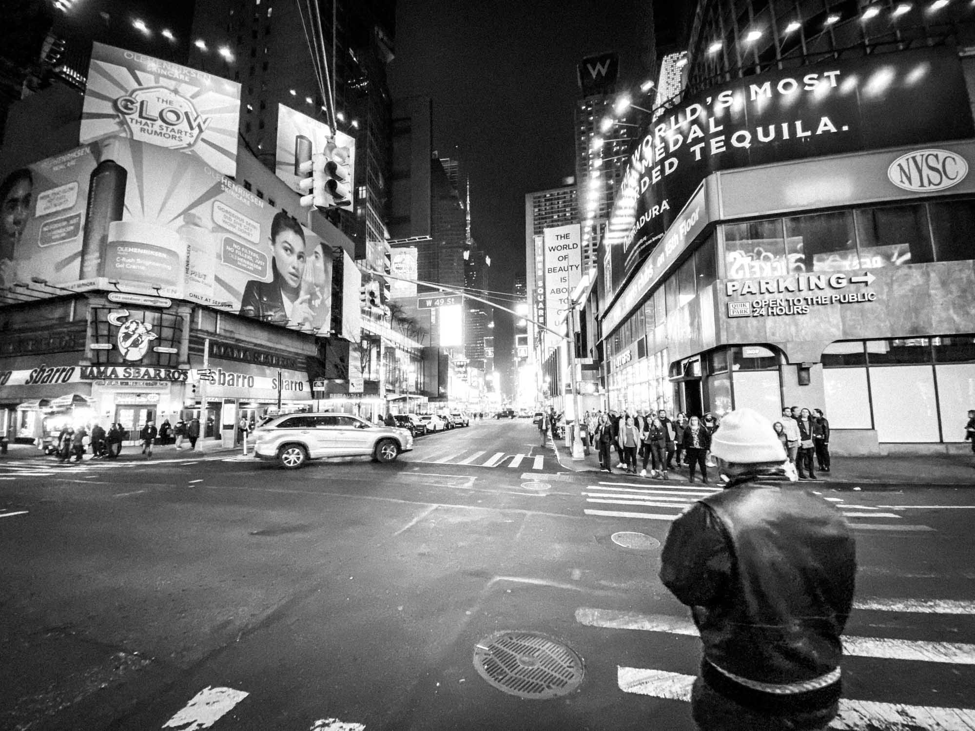 Black and white city street scene at night with bright billboards, pedestrians, and traffic at an urban intersection.