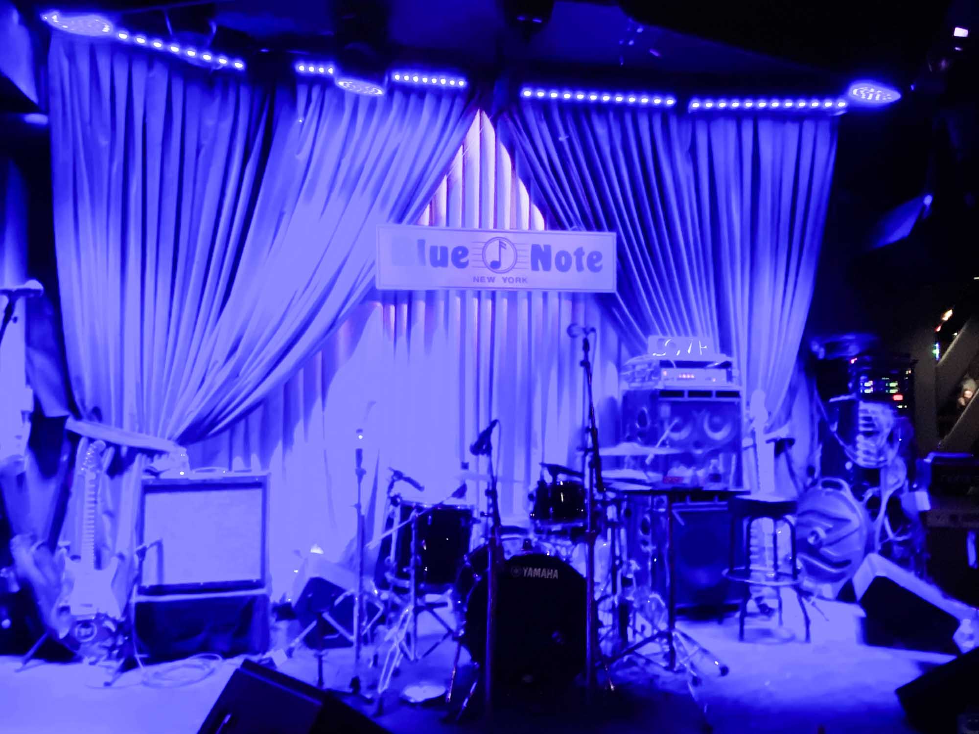 Dimly lit jazz club stage with drums, guitar, and microphone under blue lighting, Blue Note sign visible.