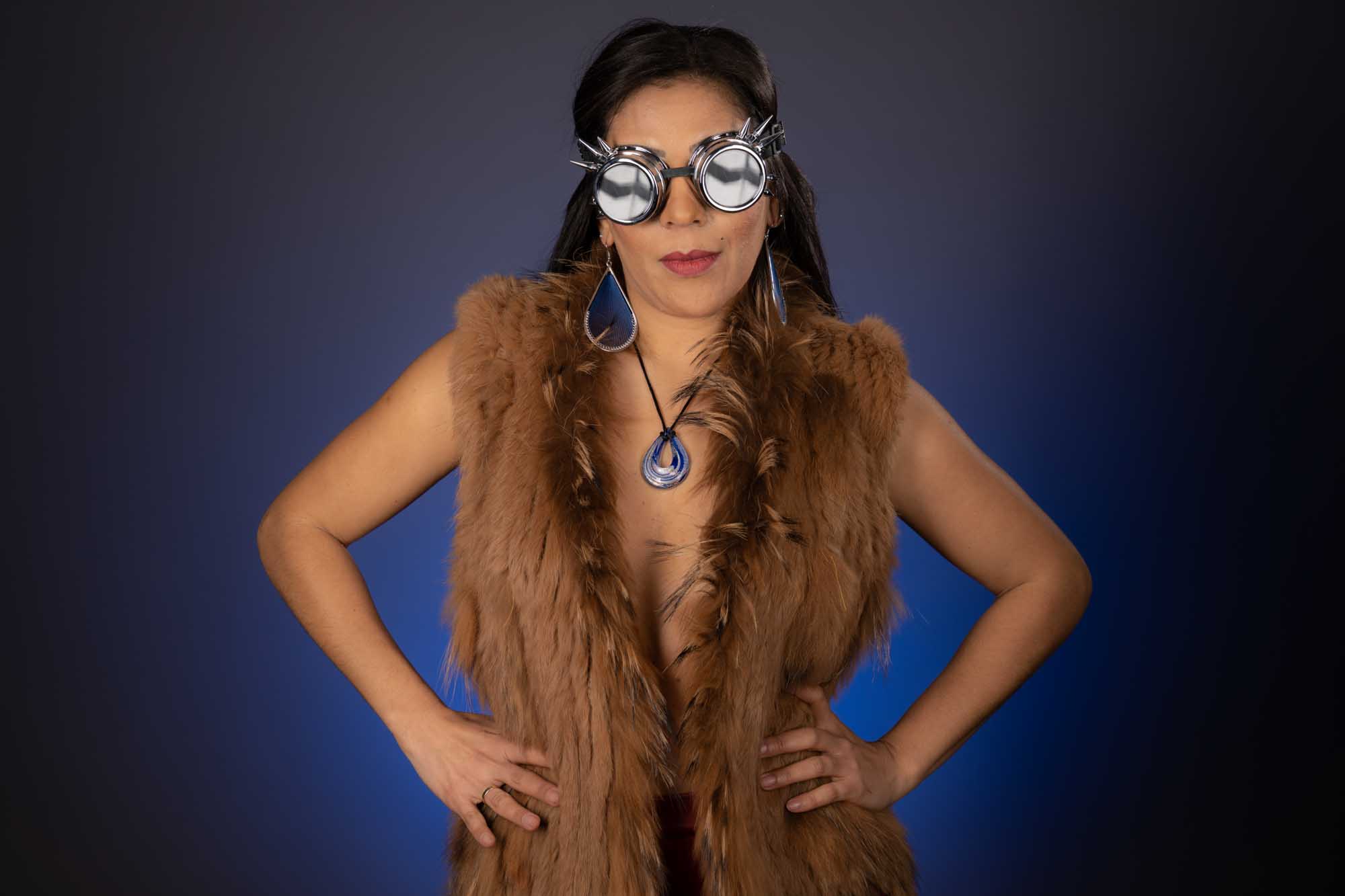 Woman in a fur vest wearing steampunk goggles, large earrings, and necklace against a dark blue background.