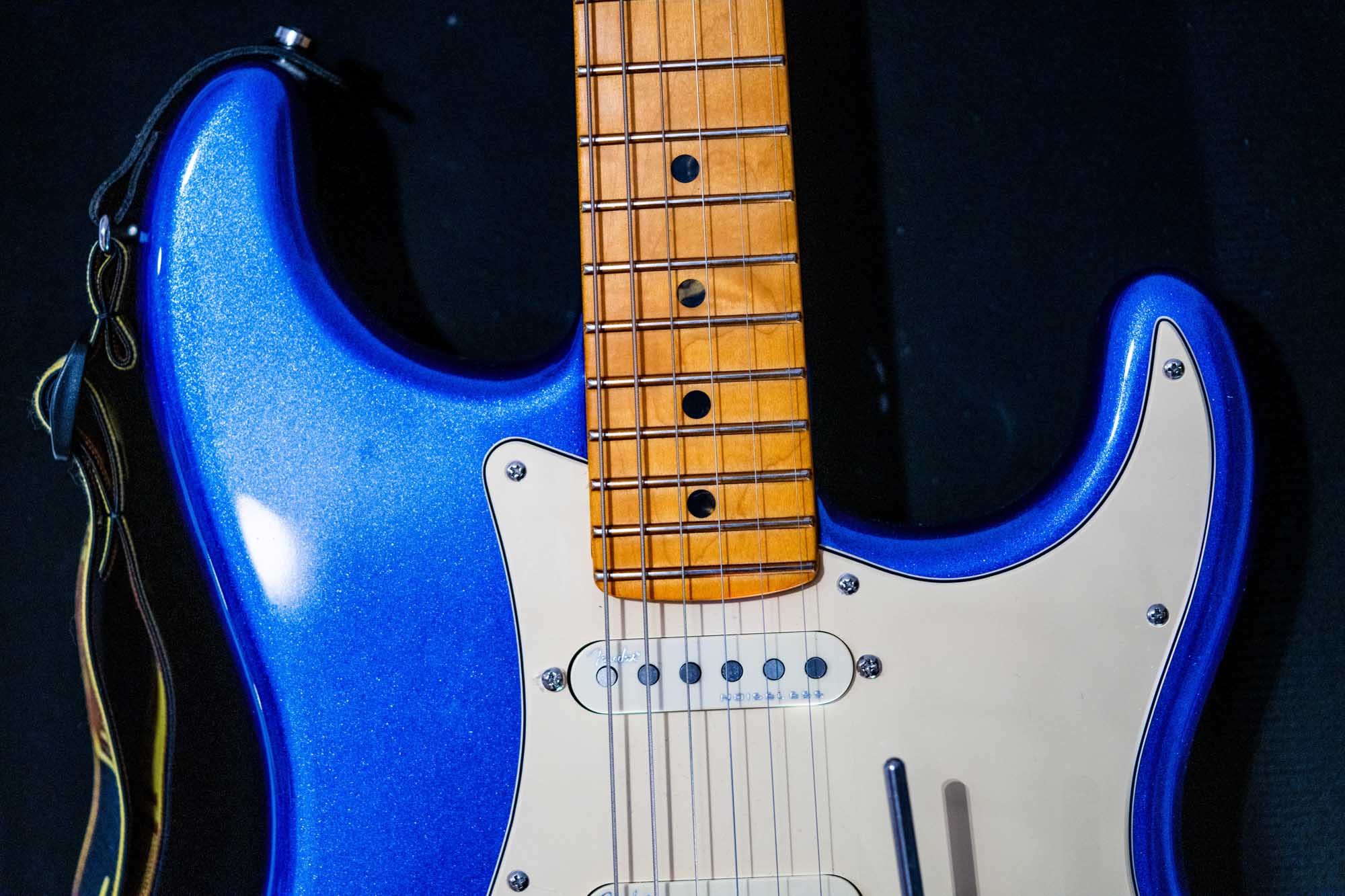 Close-up of a blue electric guitar, showing its wooden fretboard and white pickguard against a black background.