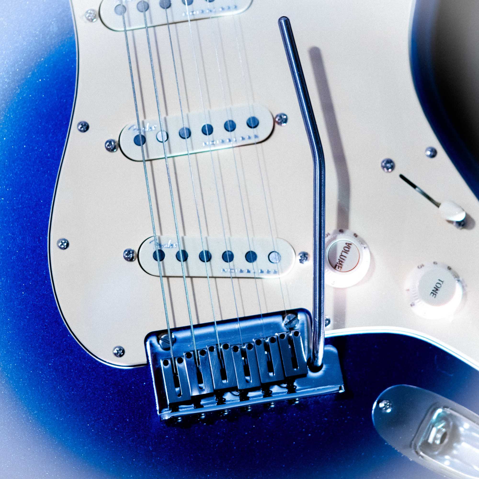 Close-up of electric guitar body with blue finish, featuring pickups, volume knobs, and a vibrato bar.
