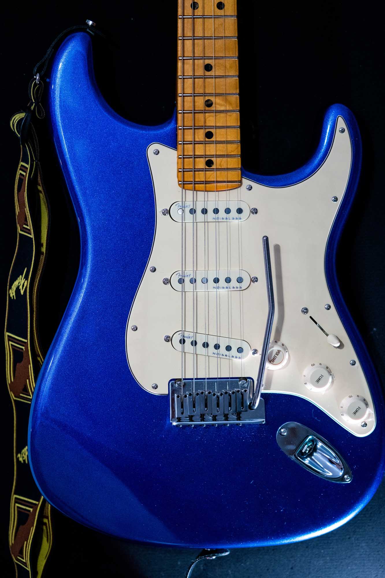 Close-up of a blue electric guitar with a white pickguard and a patterned strap against a dark background.