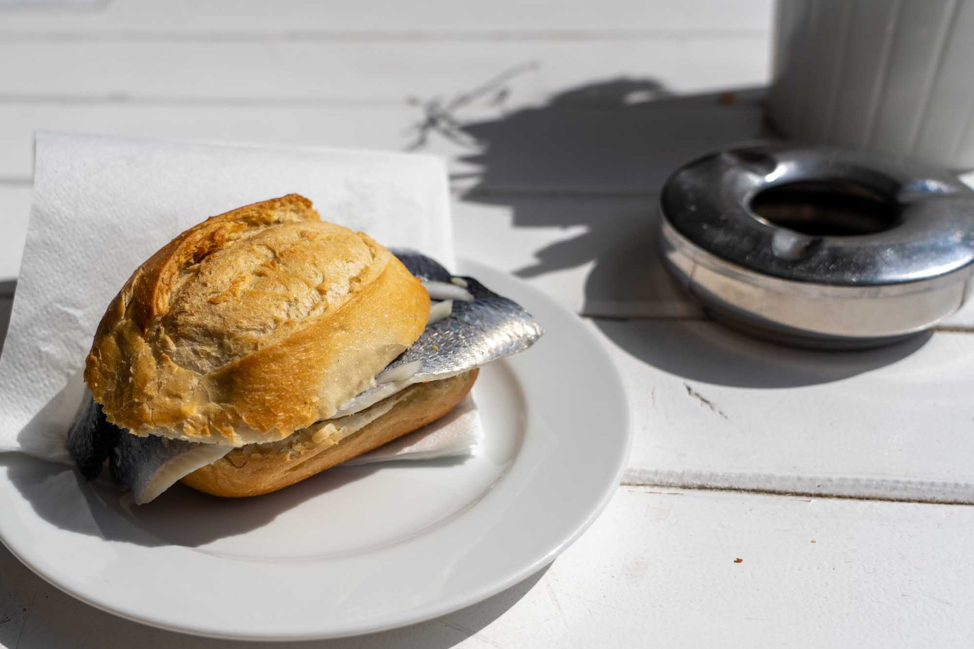 Fish roll with herring fillets on white plate outdoors, next to metallic ashtray on sunny day.