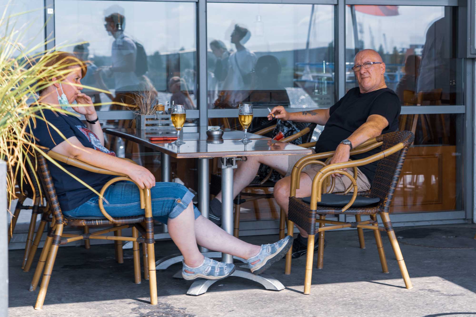 Two people seated at an outdoor café table, enjoying drinks and smoking on a sunny day.