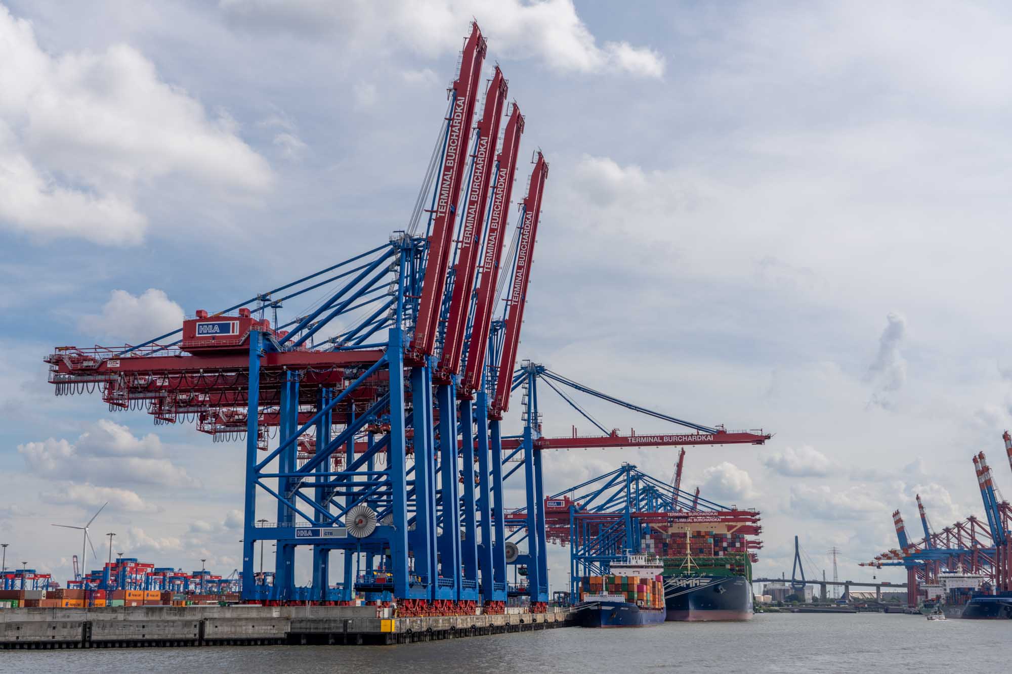 Large container cranes at a busy shipping port with cargo ships and stacked containers under a cloudy sky.