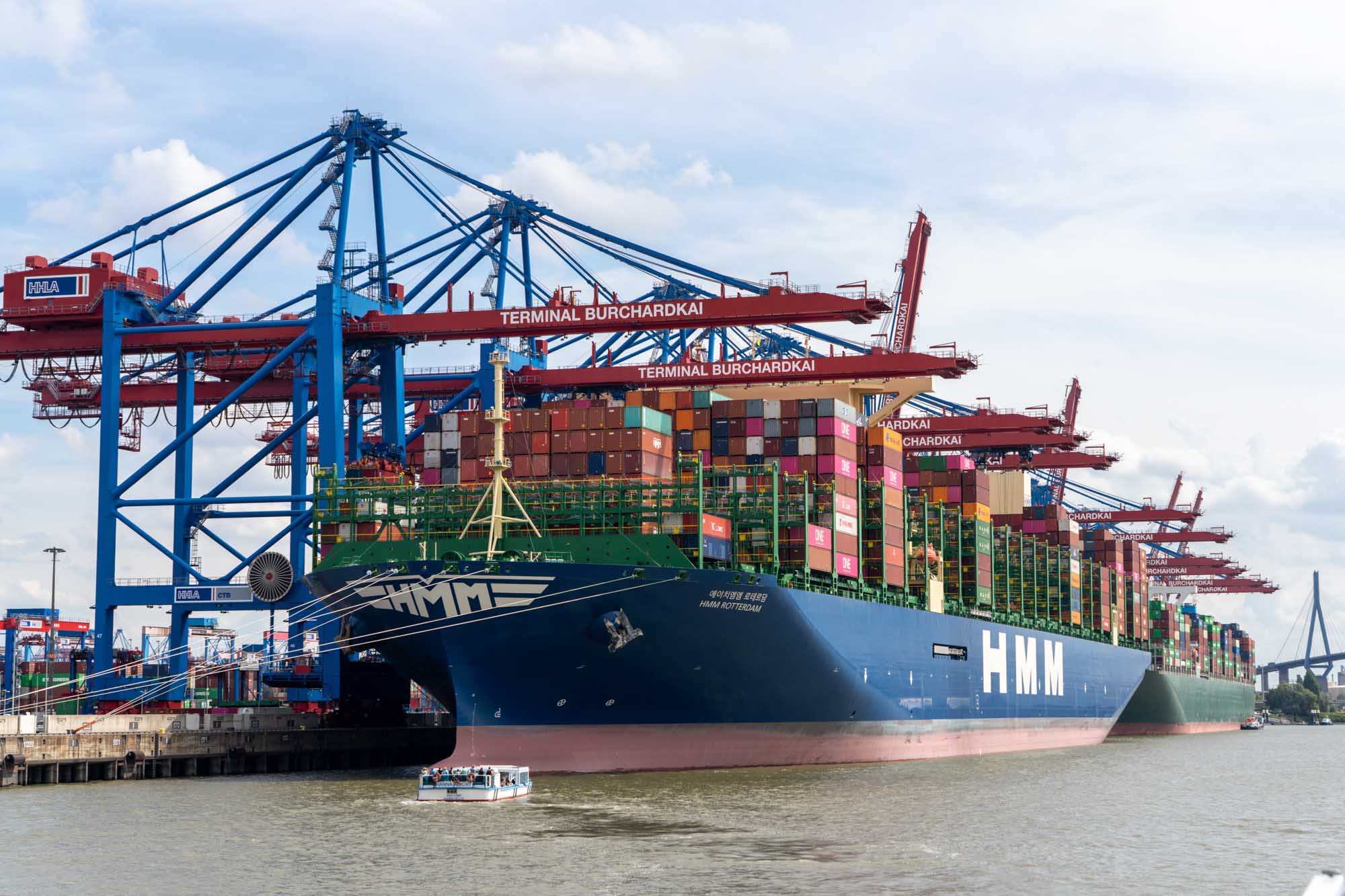 Container ship HMM Rotterdam docked at port with cranes and stacked containers, small boat in foreground.