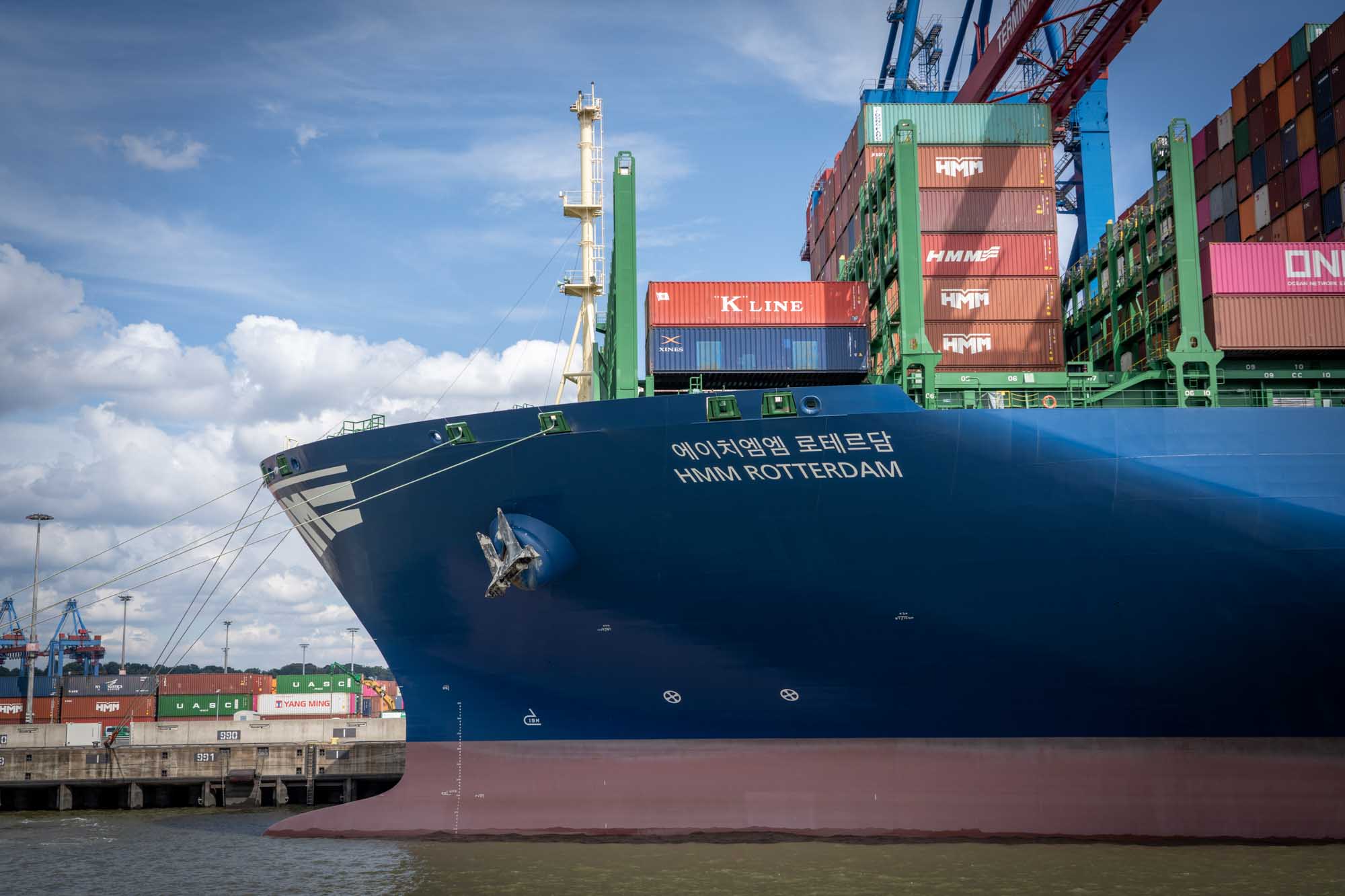 Large cargo ship HMM Rotterdam at port loaded with colorful shipping containers under a partly cloudy sky.