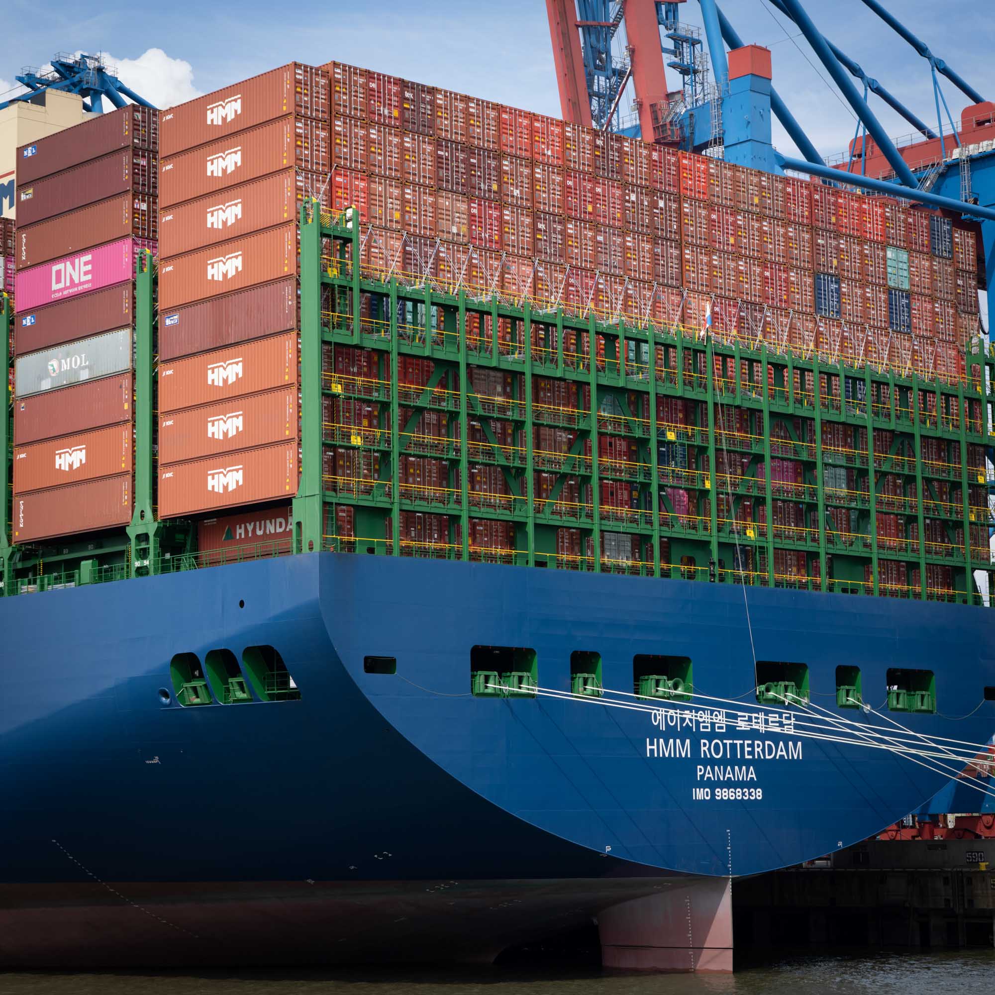 Large container ship docked, loaded high with colorful shipping containers, labeled HMM Rotterdam, in a busy port.