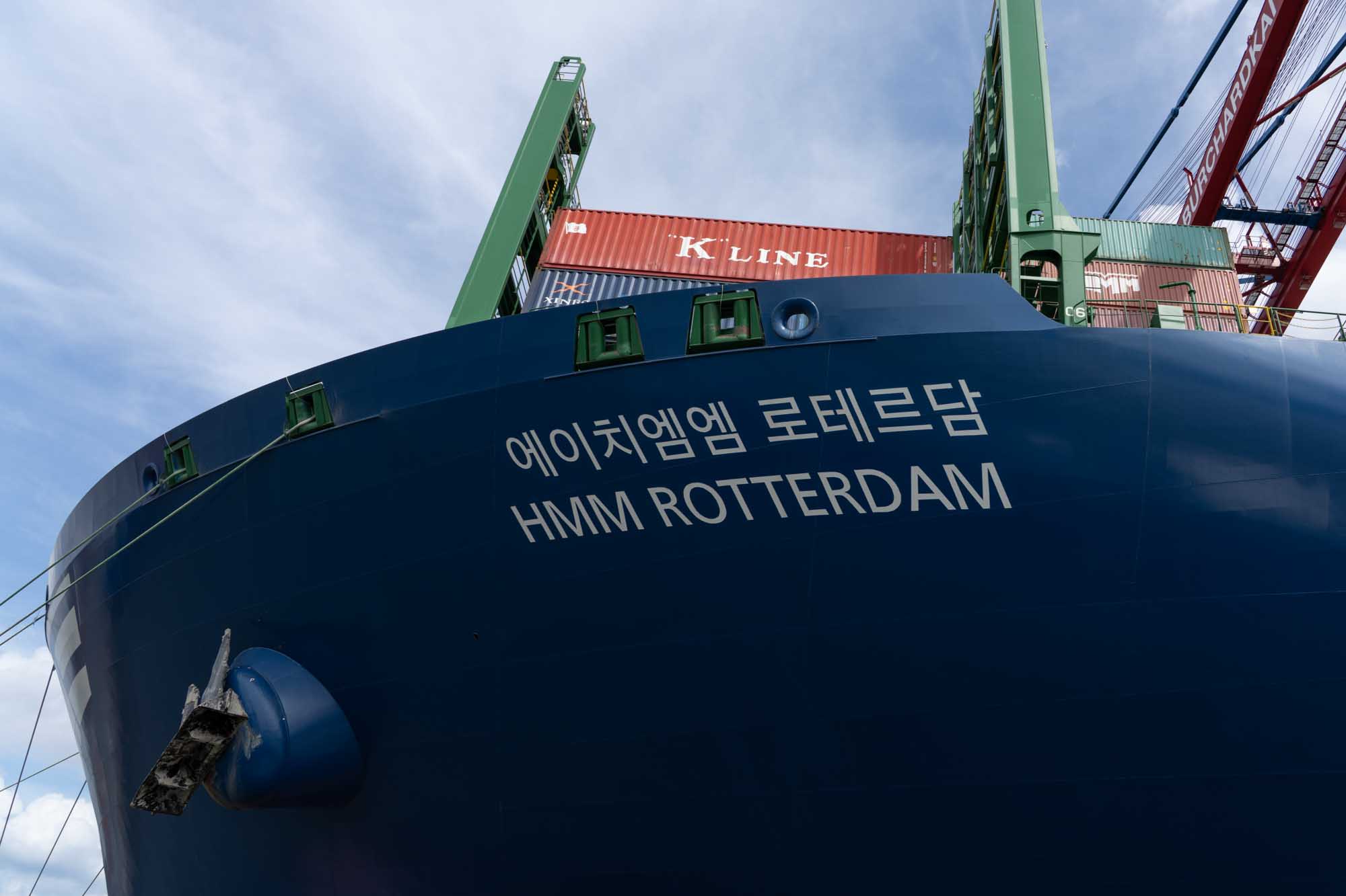 Container ship HMM Rotterdam with visible Korean text, docked under a clear sky with stacked cargo containers.
