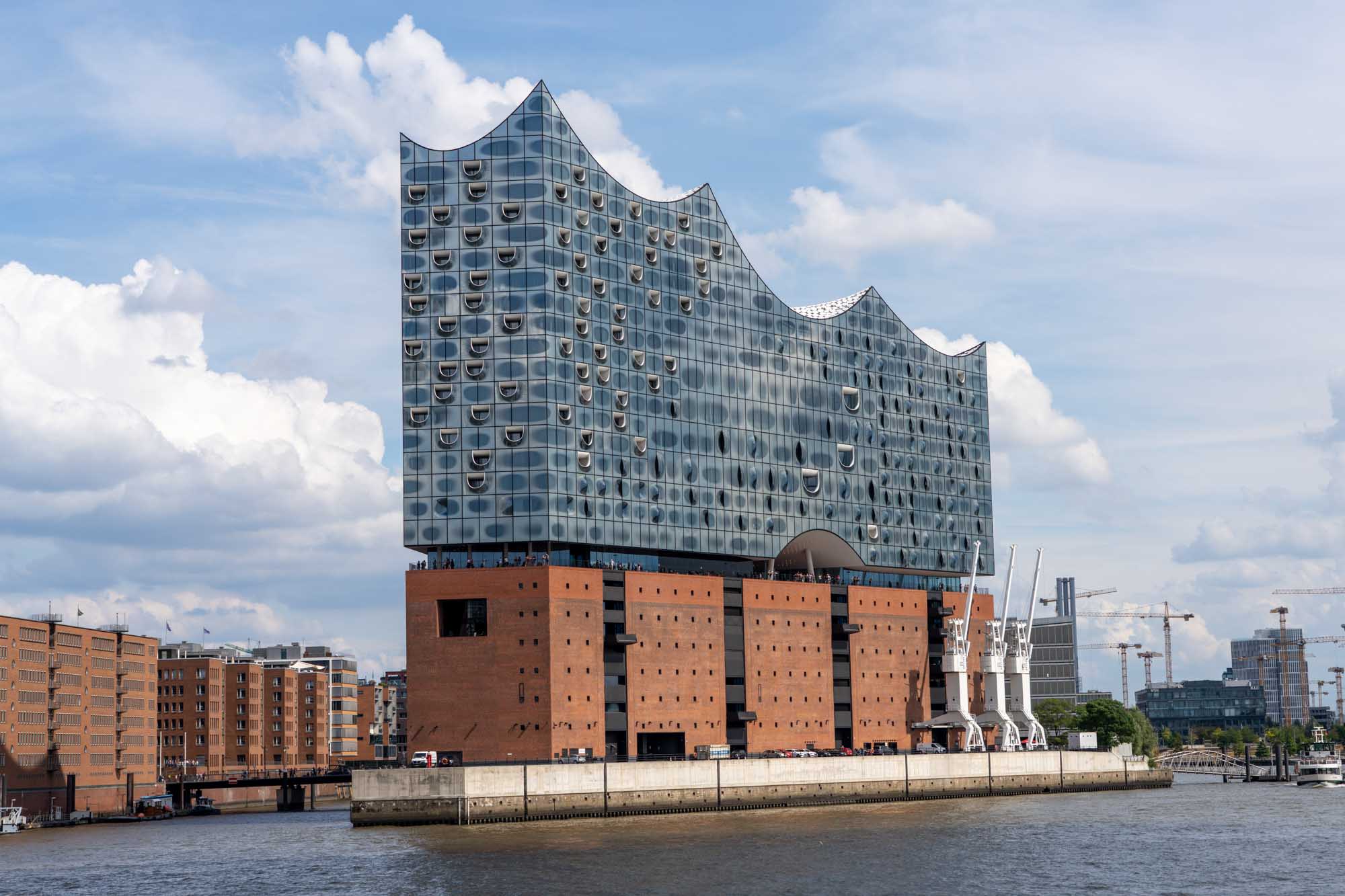 Modern waterfront building with a wavy glass facade and brick base against a cloudy sky, near construction cranes.